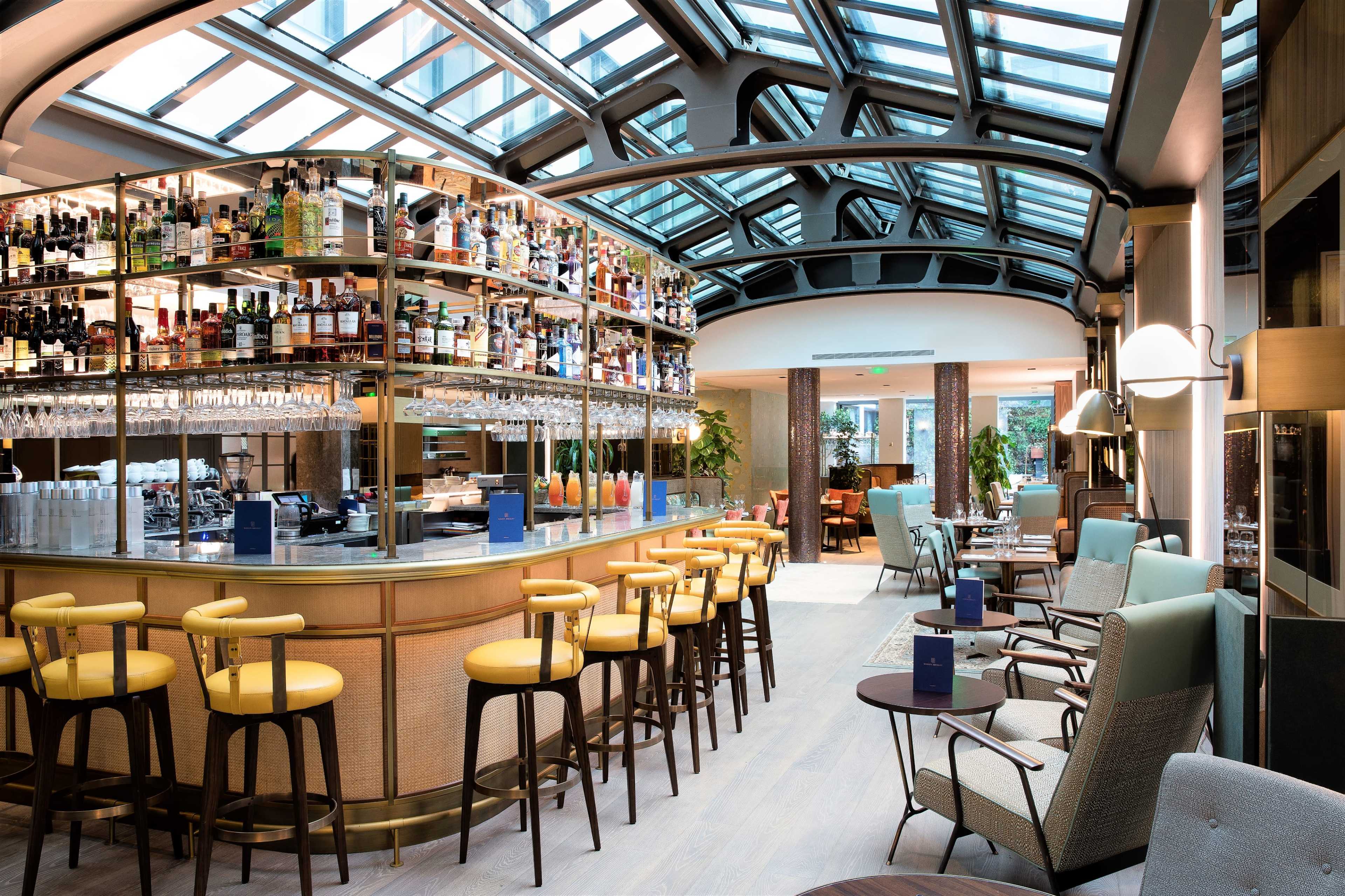 A modern bar area inside a well-lit restaurant, featuring a glass ceiling and shelves stocked with various bottles.