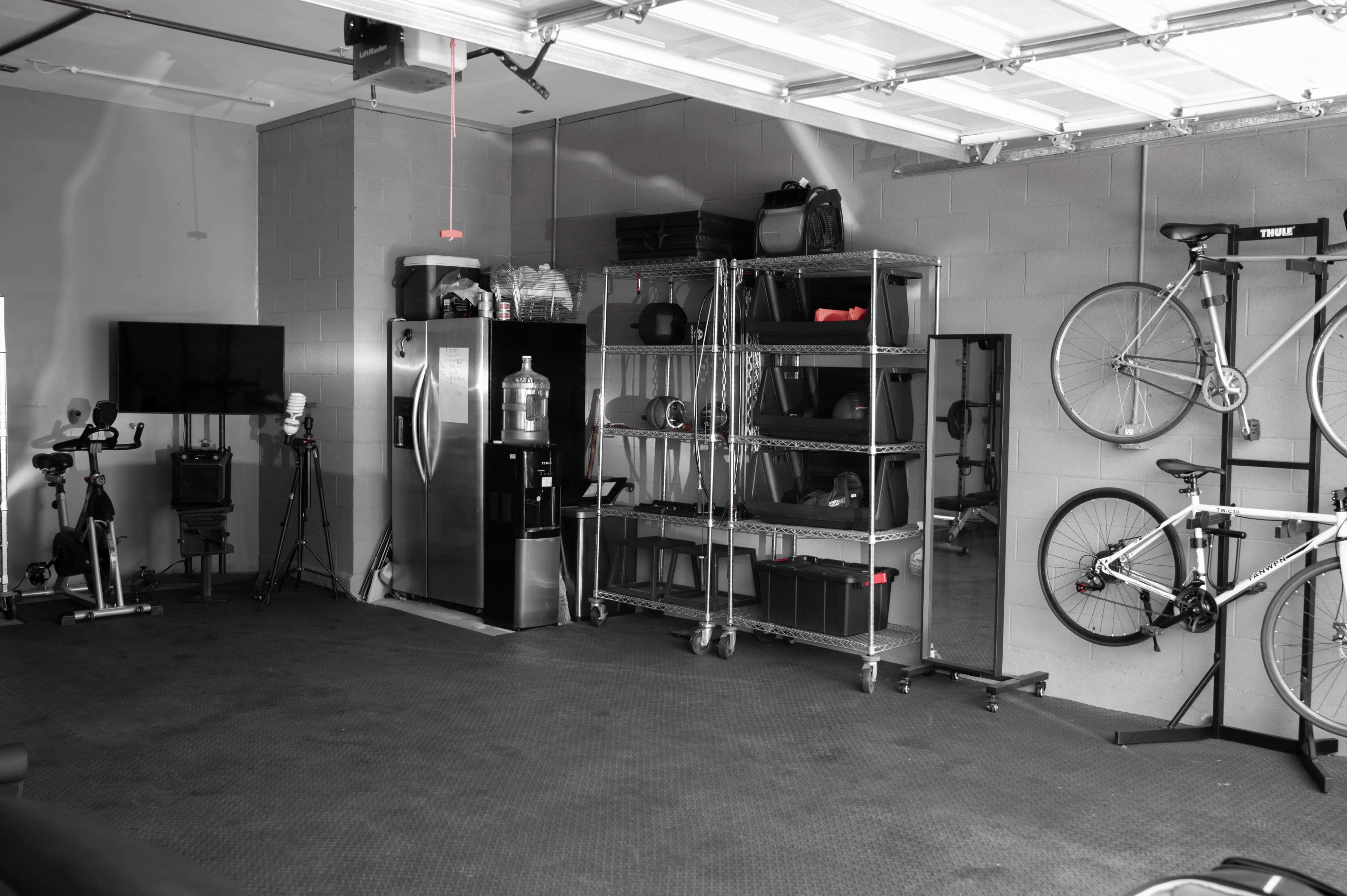 A spacious garage features a refrigerator, a water cooler, a shelving unit with various items, a stationary bike, and two bicycles mounted on the wall.
