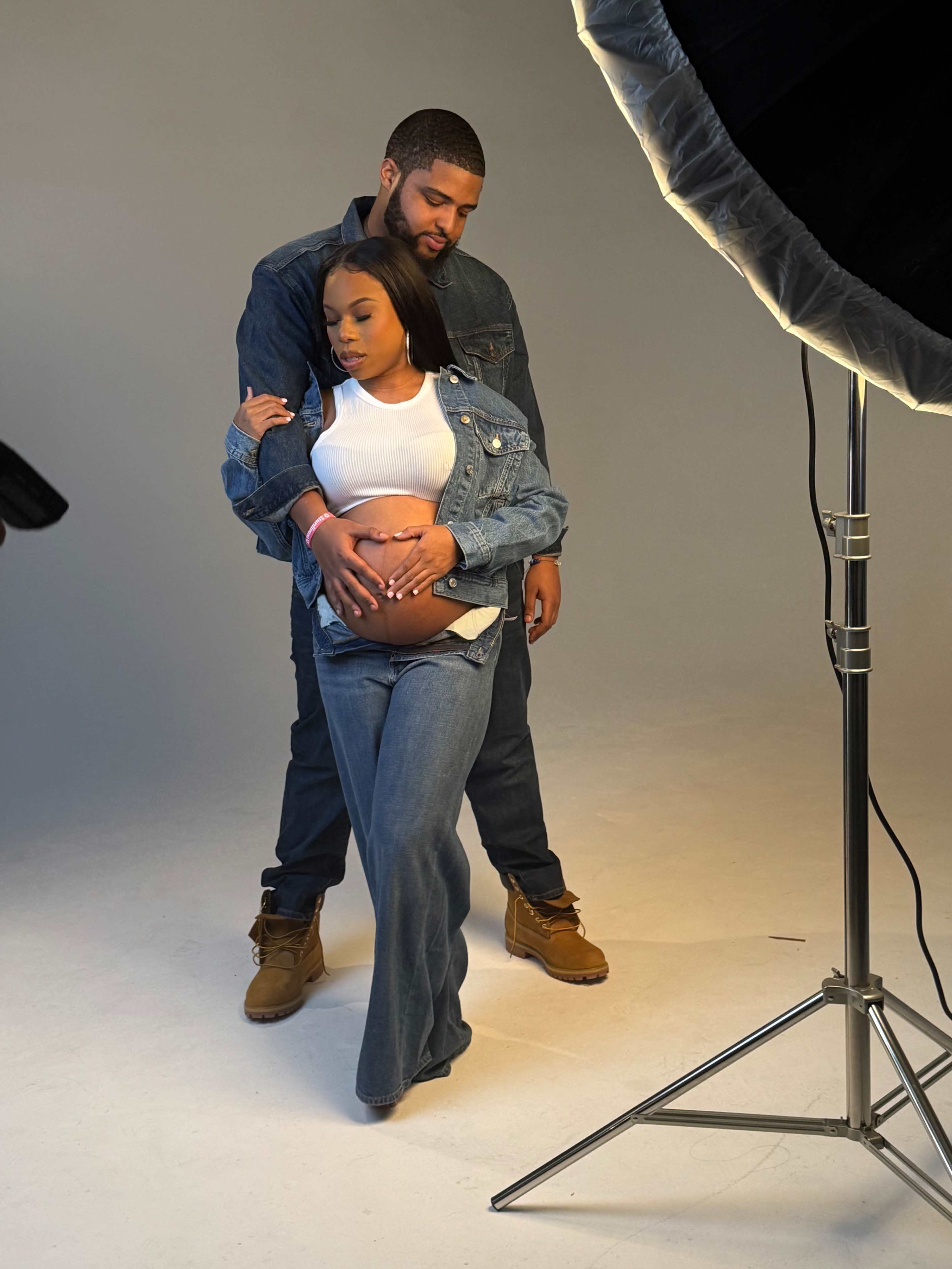 A couple poses in a studio setting, with the woman gently cradling her baby bump while wearing denim, and the man stands behind her in a matching outfit.