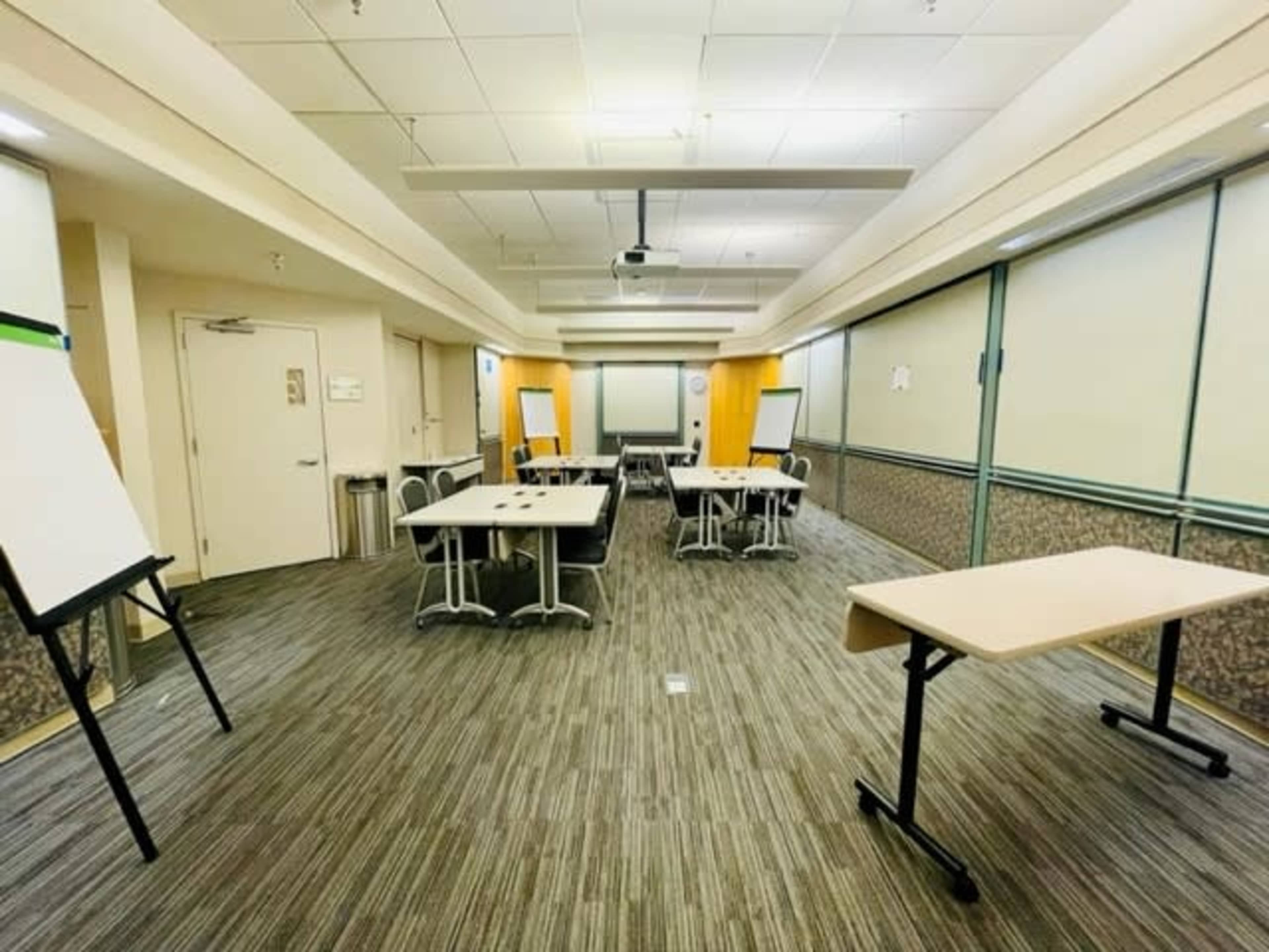 A conference room with several tables and chairs arranged for a meeting, featuring whiteboards and a projector.