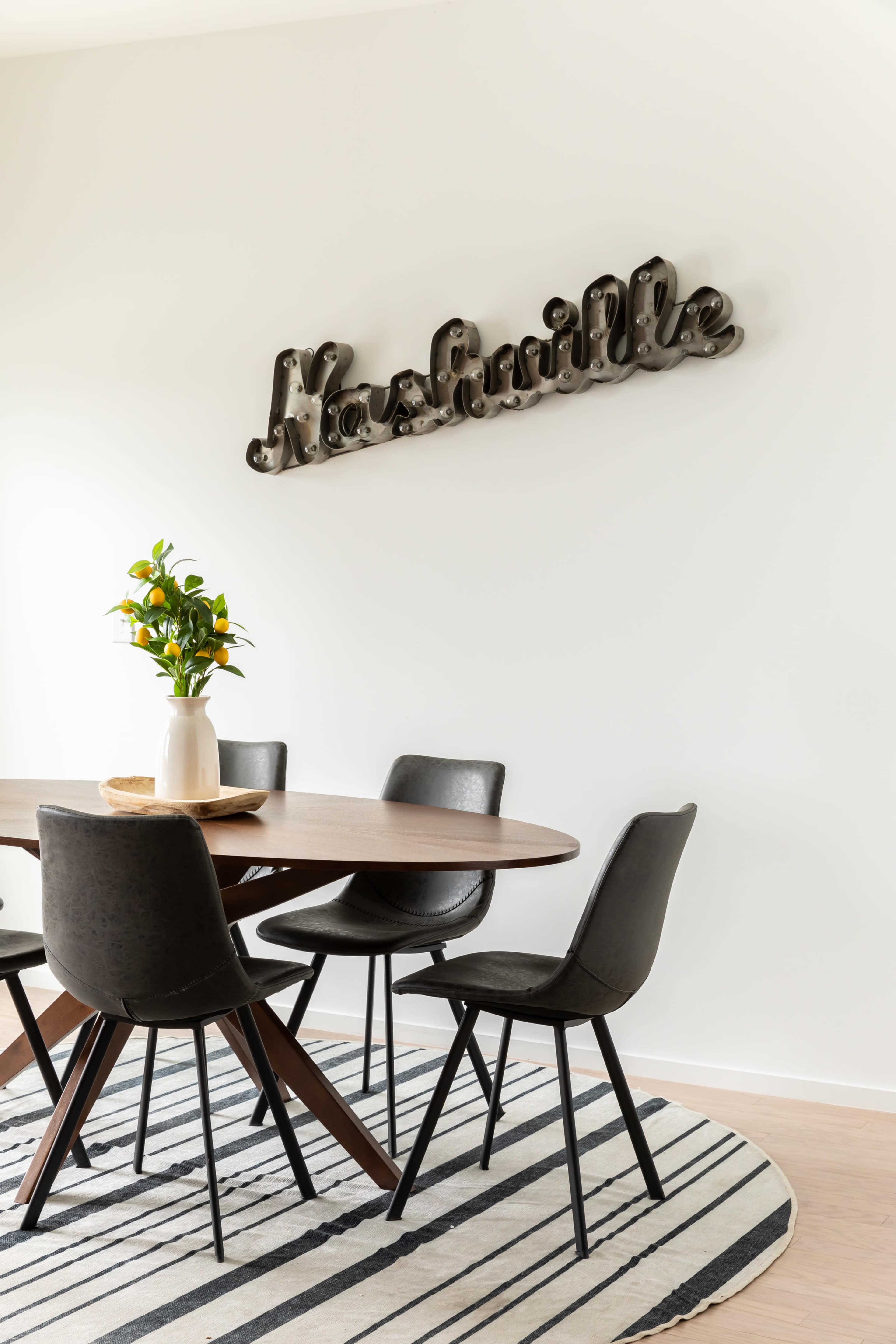 A round wooden dining table is surrounded by black chairs under a decorative "Nashville" sign mounted on the wall, with a vase of flowers in the center.