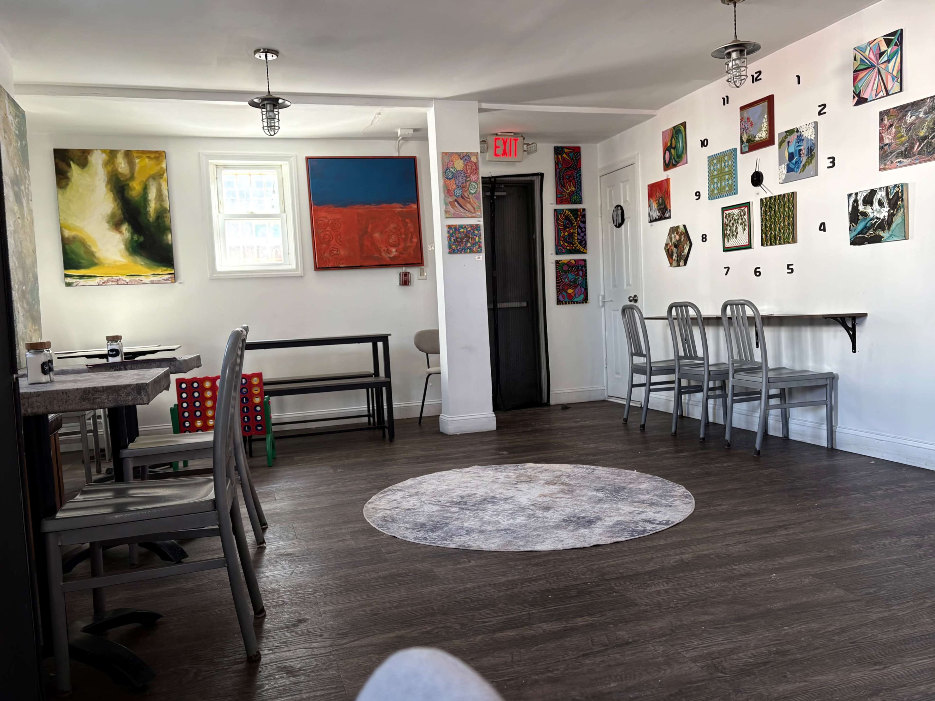 The image shows an art-filled café with several tables and chairs, a circular rug on the floor, and colorful paintings displayed on the walls.