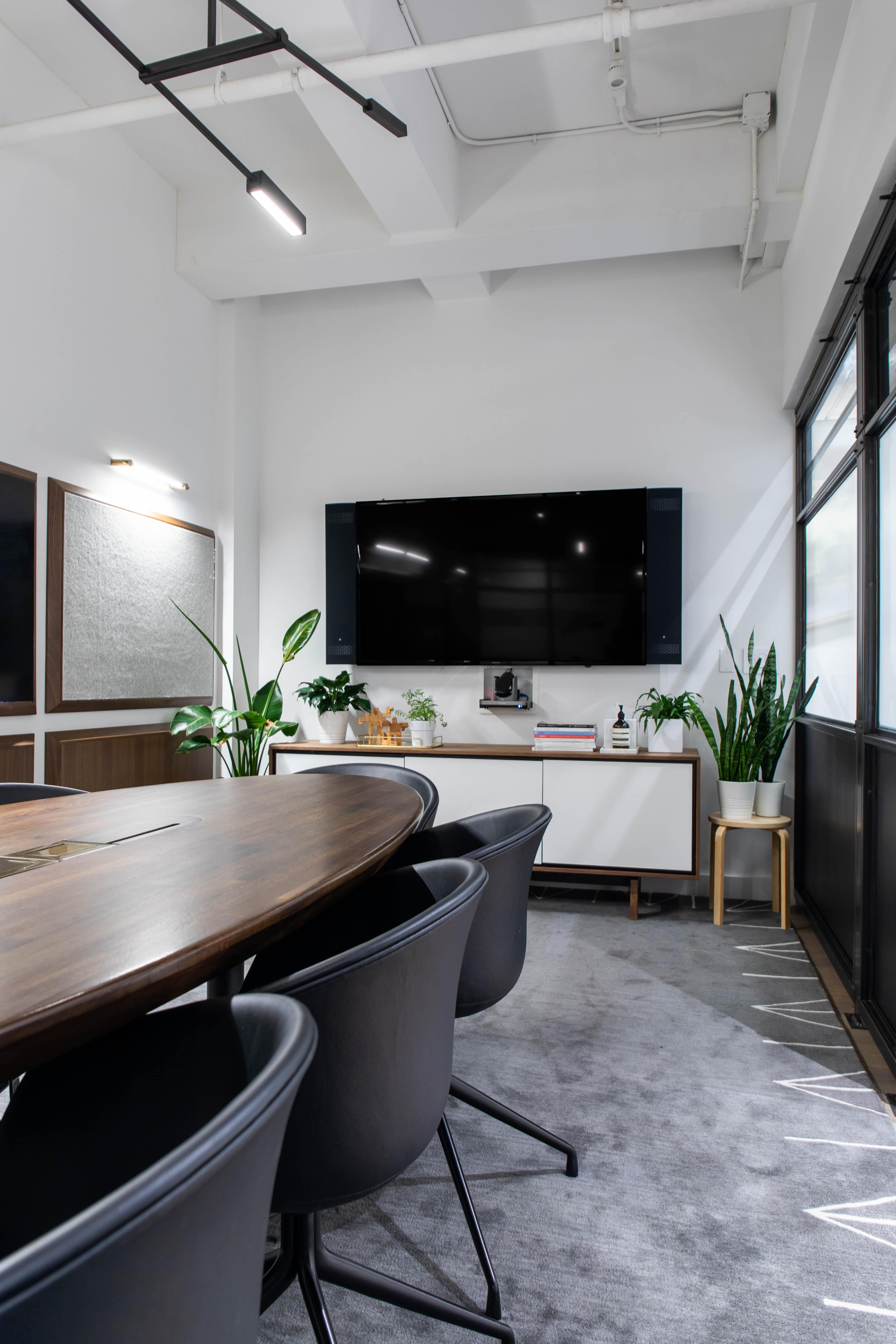 A modern conference room features a large wooden table, black chairs, a television mounted on the wall, and several potted plants.