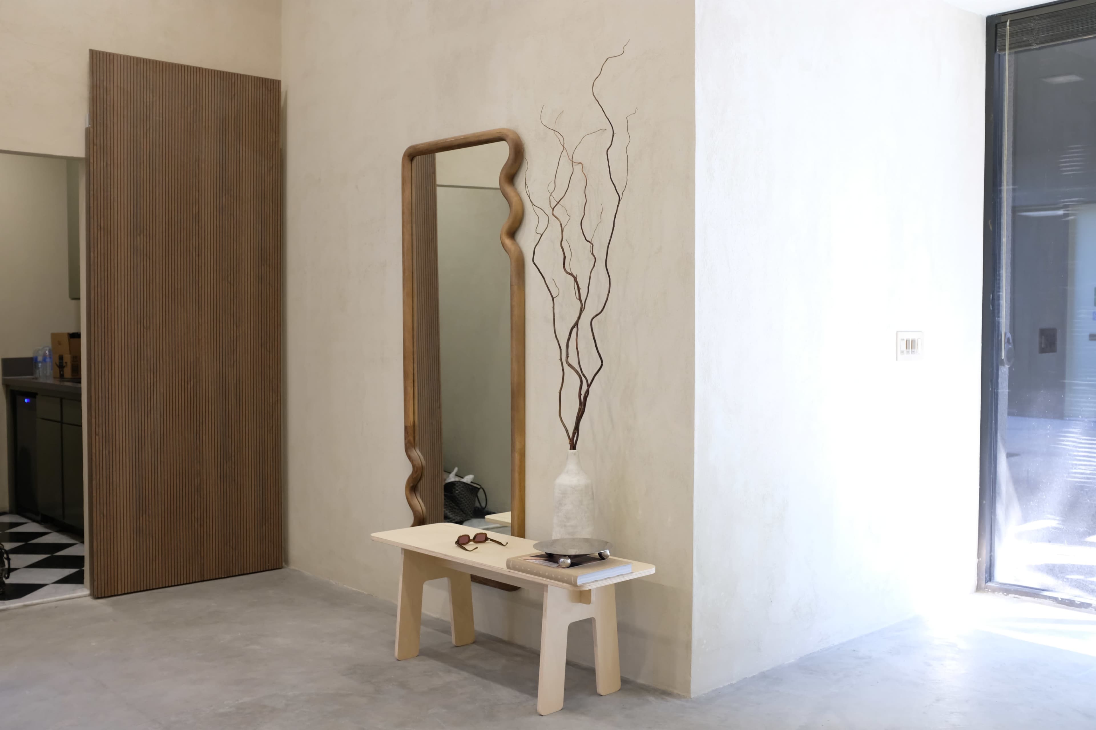 The image shows a minimalist entryway featuring a wooden mirror, a simple bench, and a decorative vase with branches against a neutral wall.