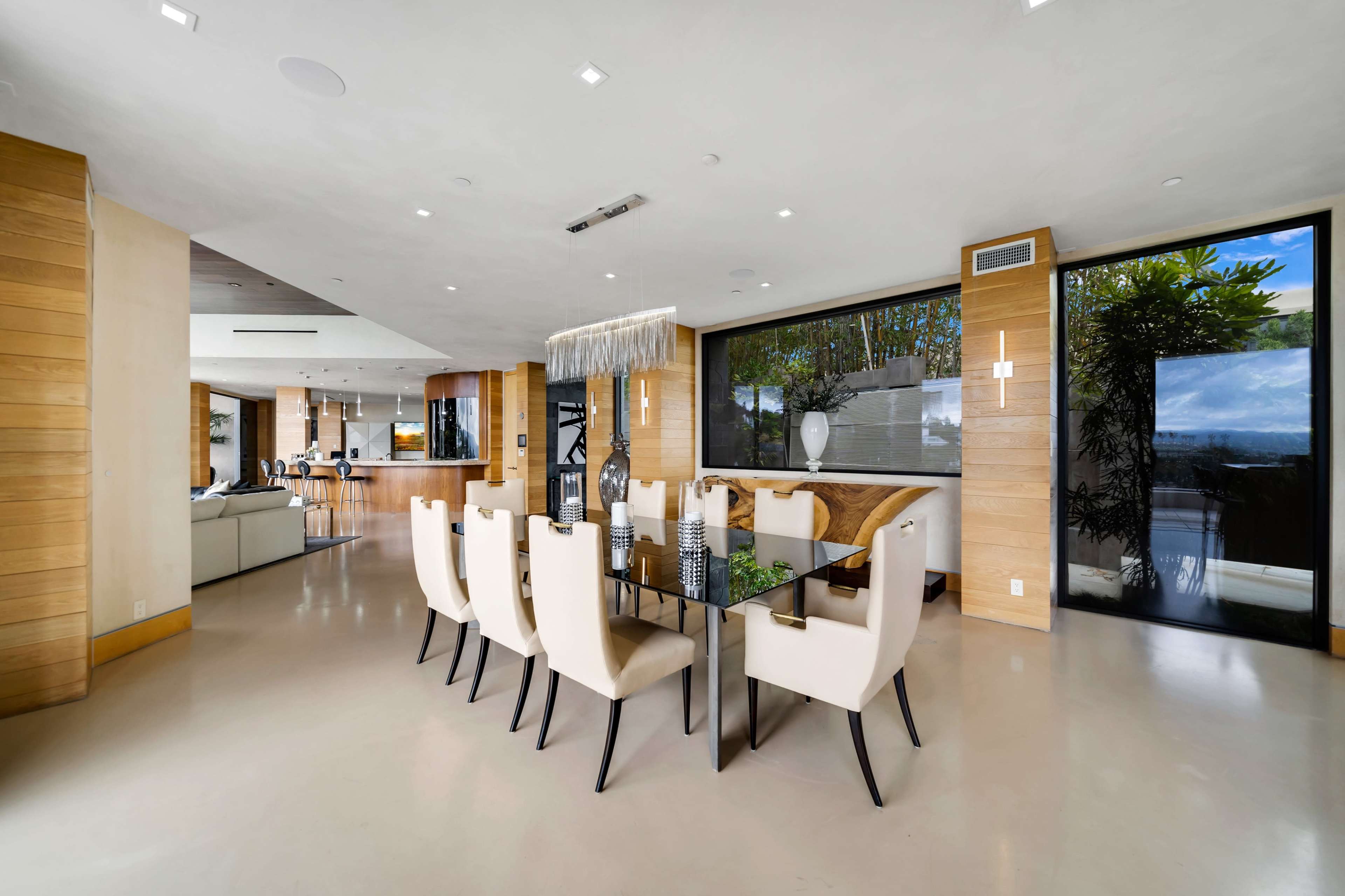 A modern dining area features a glass table surrounded by white chairs, with an open layout that connects to a bright living space and large windows showcasing outdoor greenery.