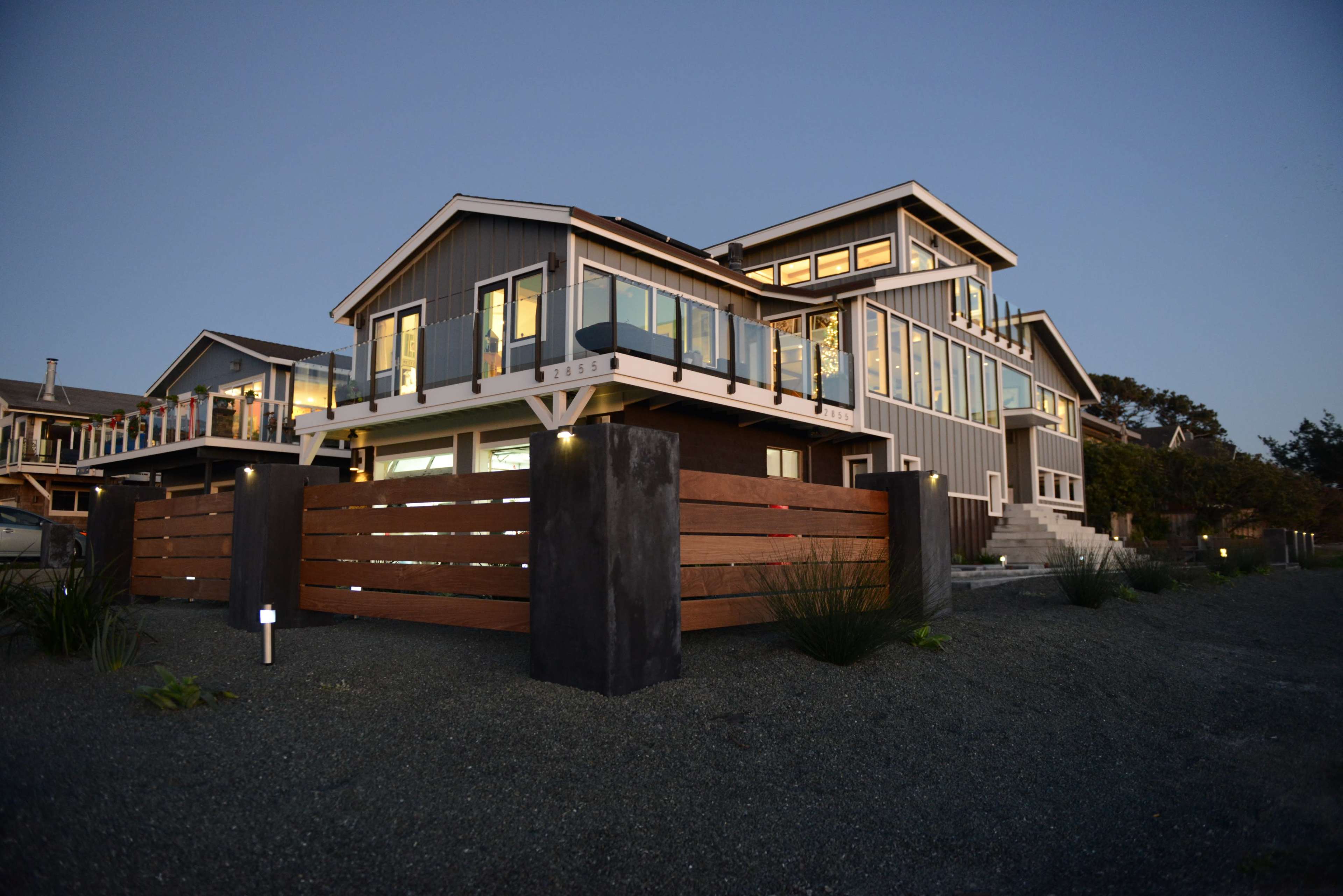 A modern coastal house with multiple levels and large windows is surrounded by a wooden fence and gravel landscape.