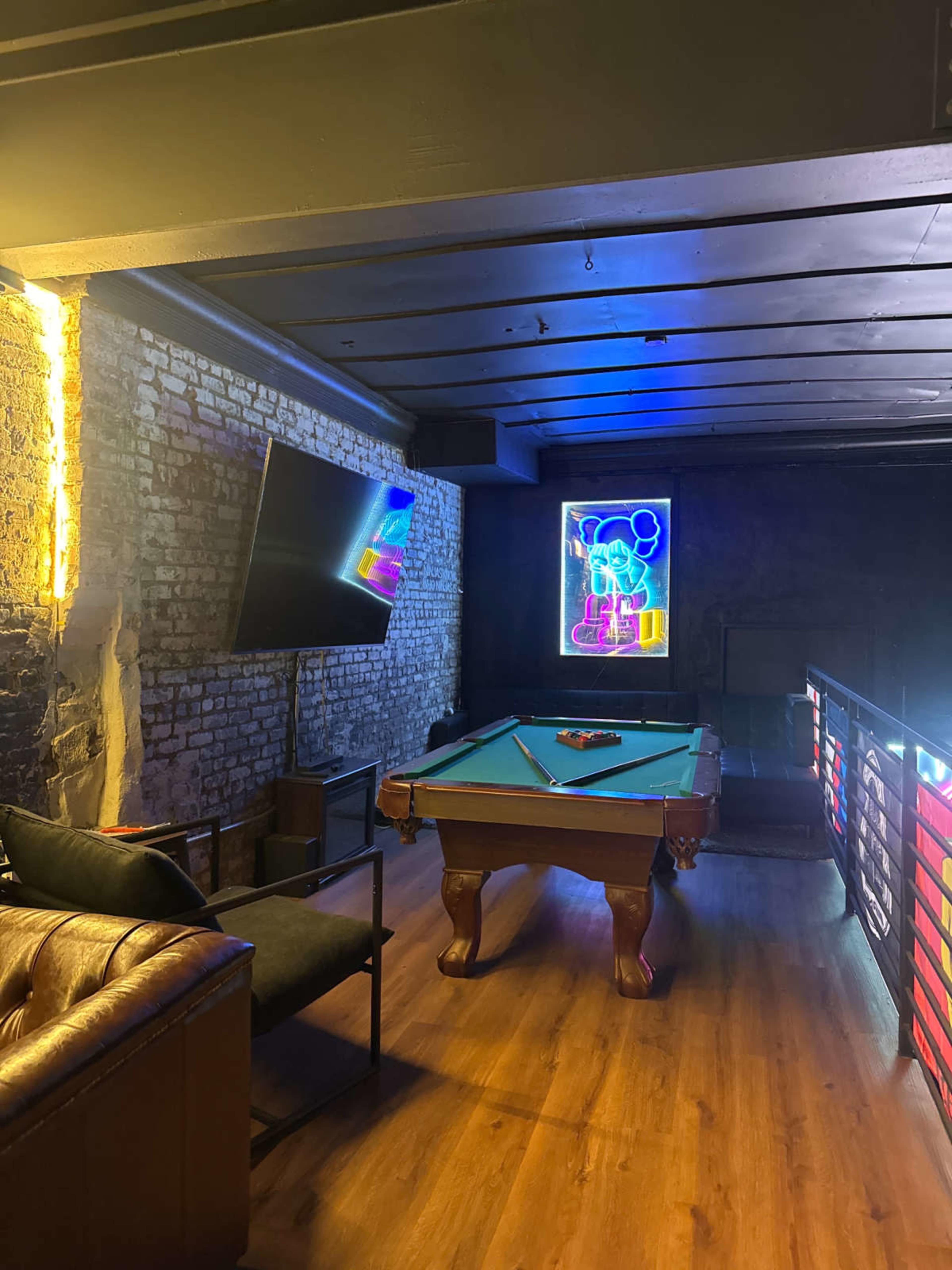 The image shows a dimly lit lounge area featuring a pool table, a large television mounted on the wall, and a colorful neon artwork.