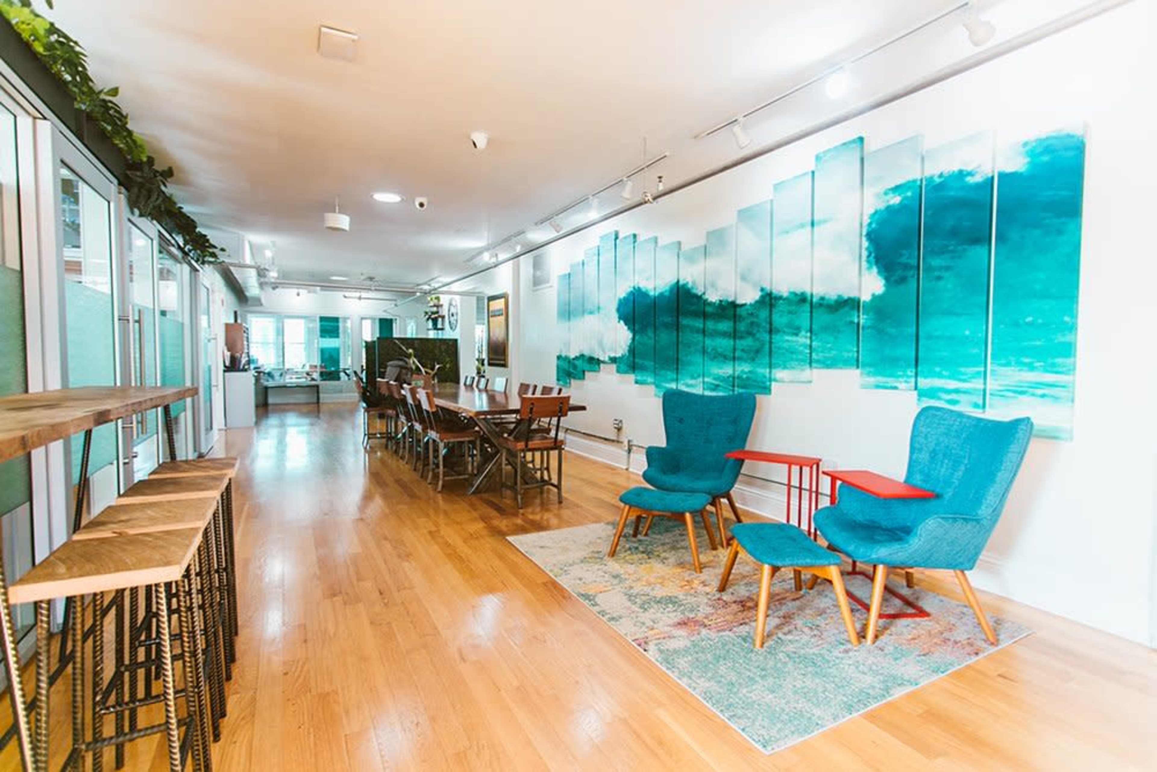 The image shows a modern office space with wooden flooring, a long communal table, a series of teal artwork depicting waves on the wall, and blue chairs arranged around a small red table.