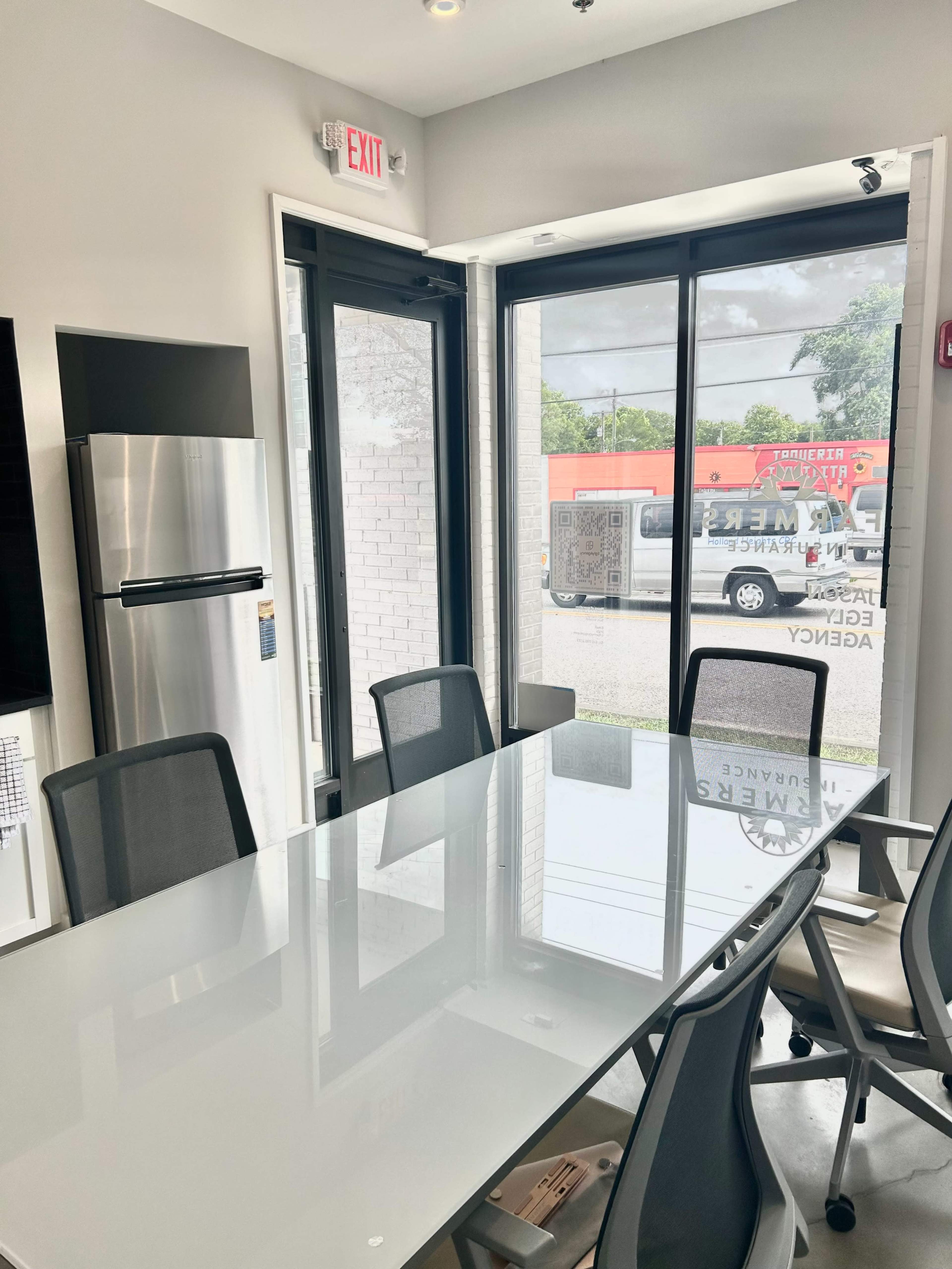 A modern conference room features a large glass table surrounded by black chairs, with a stainless steel refrigerator and large windows looking out onto a parking area.