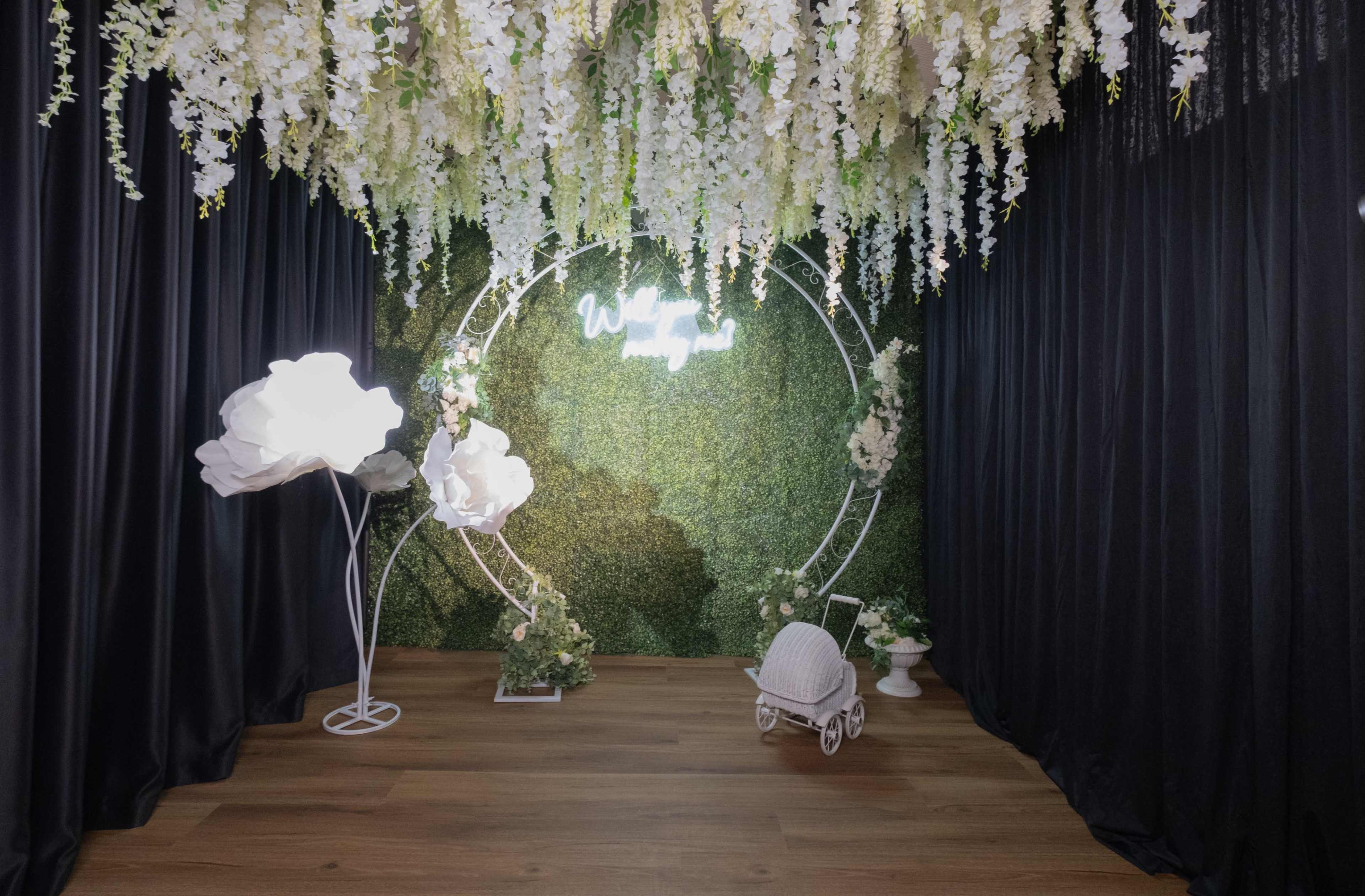 The scene features a decorative setup with a green textured backdrop, hanging floral arrangements, and a small white baby carriage on a wooden floor.