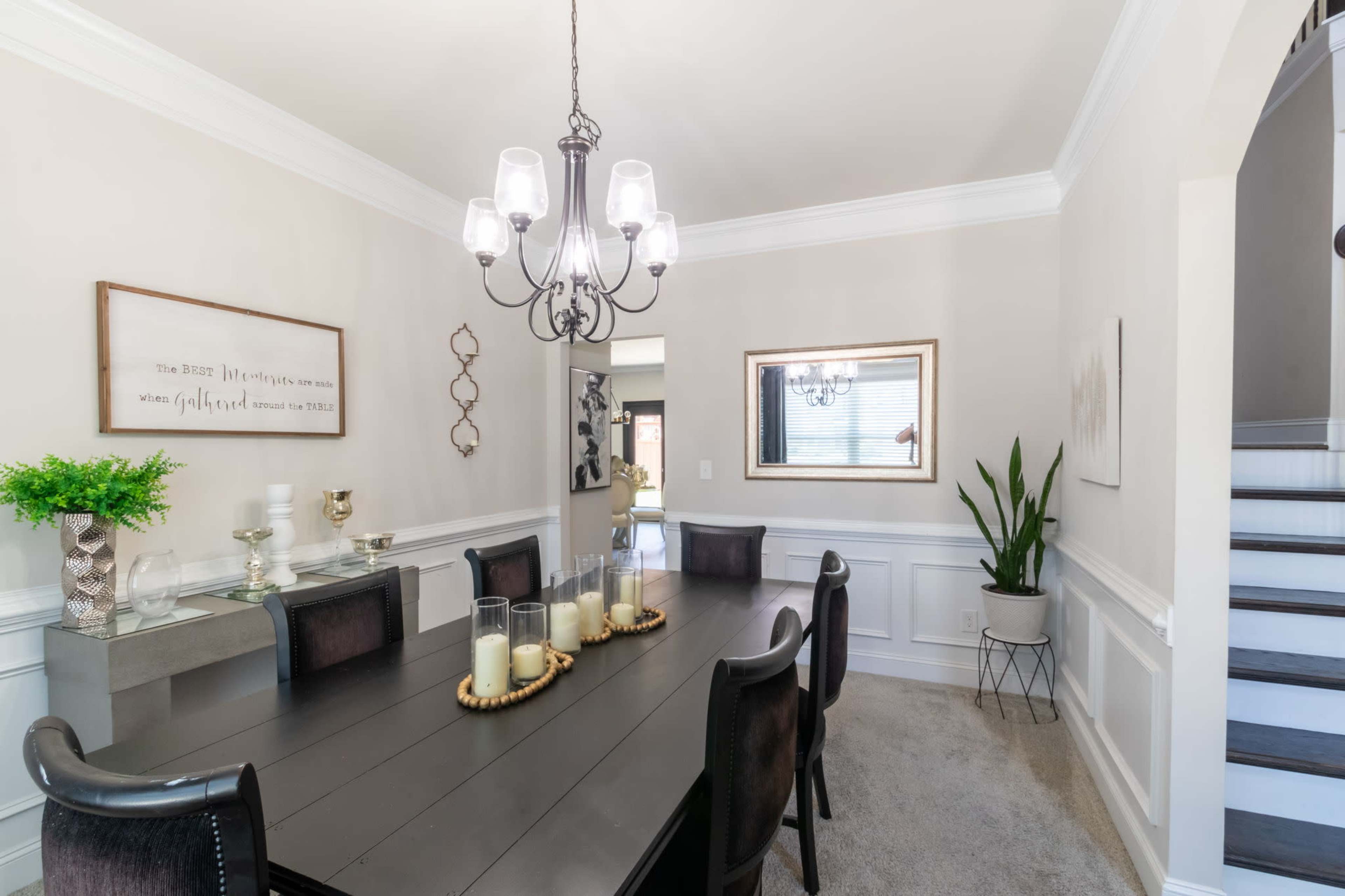 A dining room features a long black table with a centerpiece of candles, surrounded by dark chairs, and is accented by wall decor and a staircase in the background.