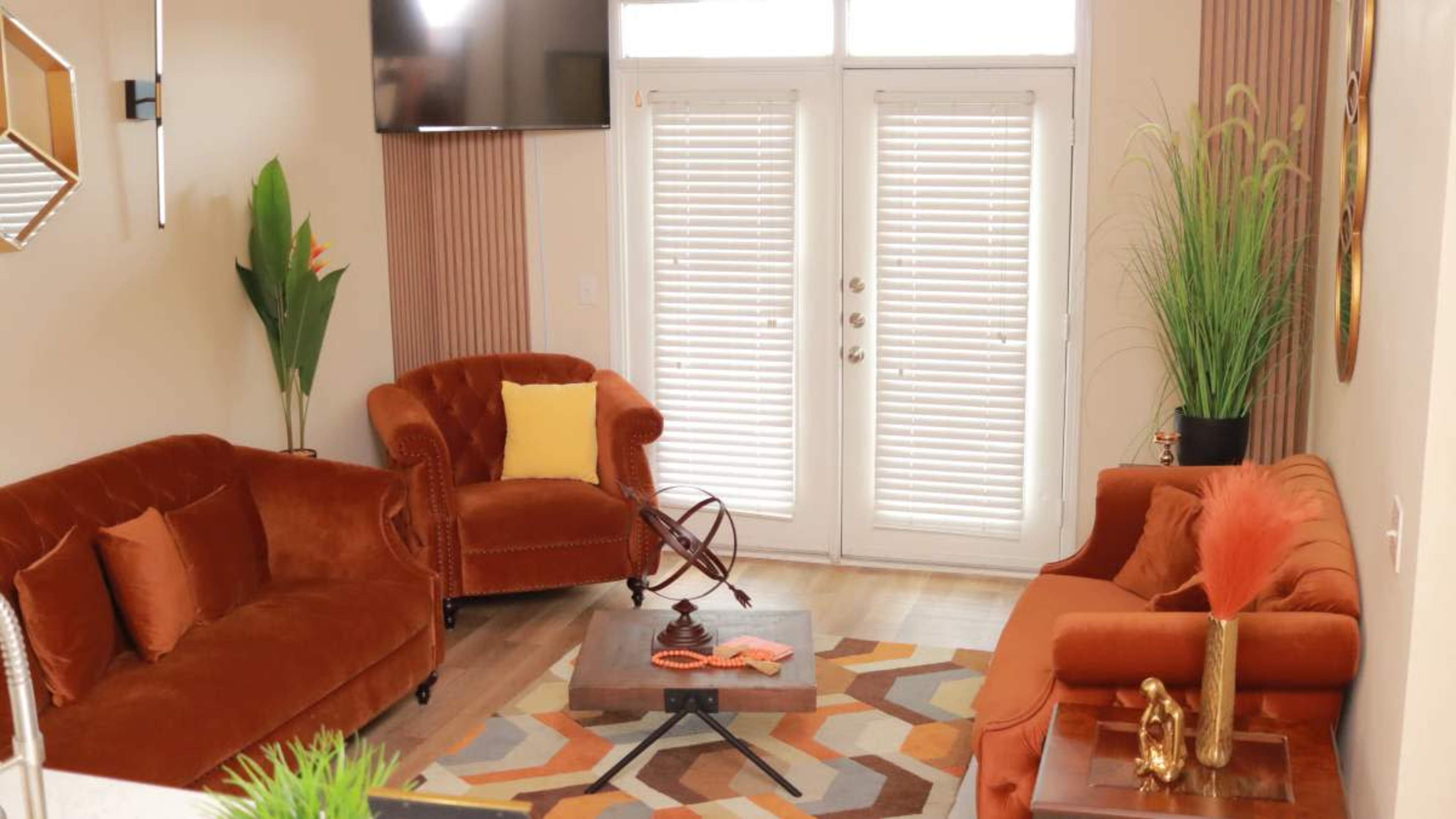 The image shows a modern living room with two orange velvet sofas, a coffee table, and large windows with closed blinds.