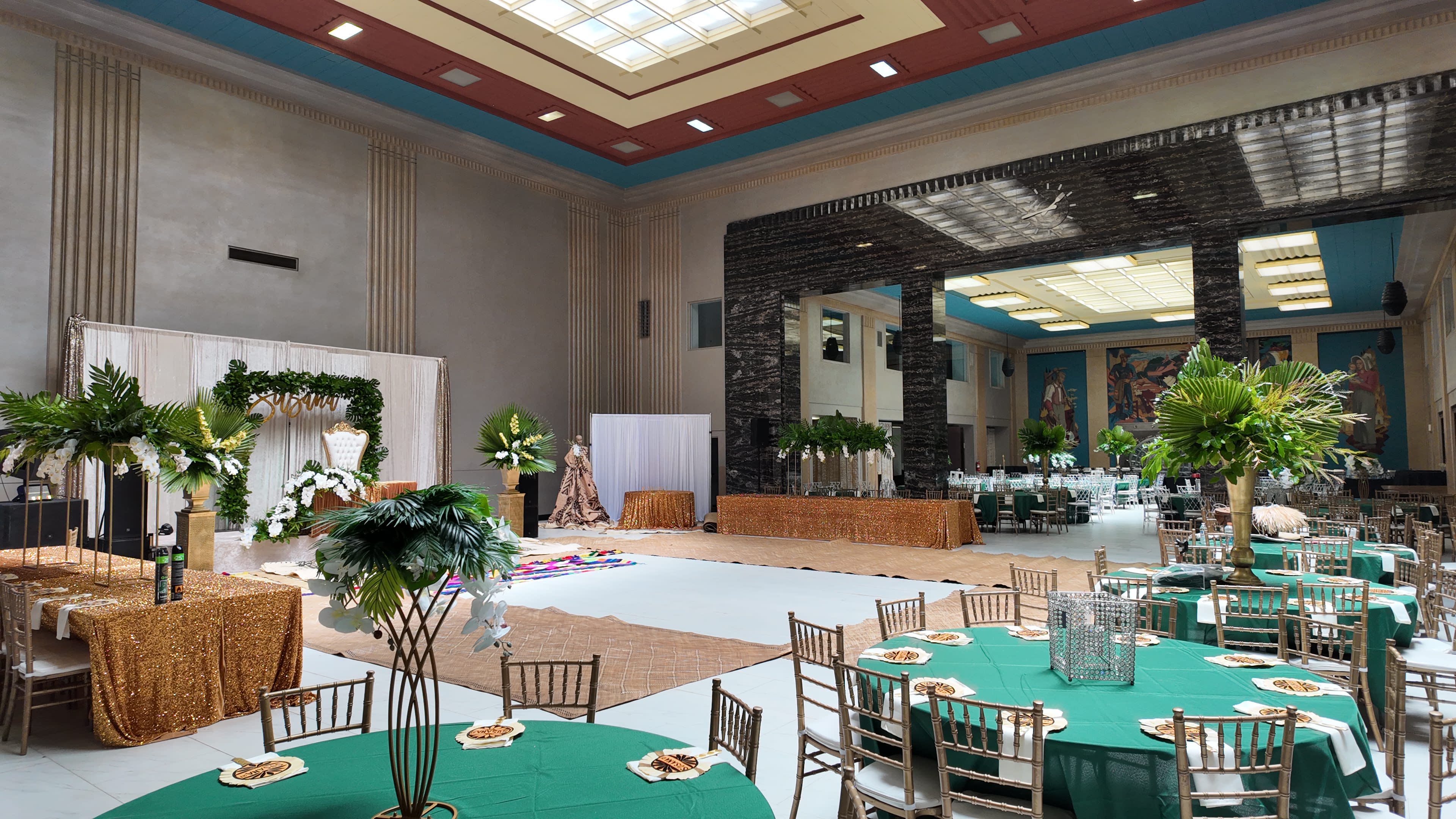 The image shows an elegantly decorated banquet hall featuring round tables covered with green tablecloths, golden accents, and large plants, with a backdrop of tall stone pillars and artwork on the walls.