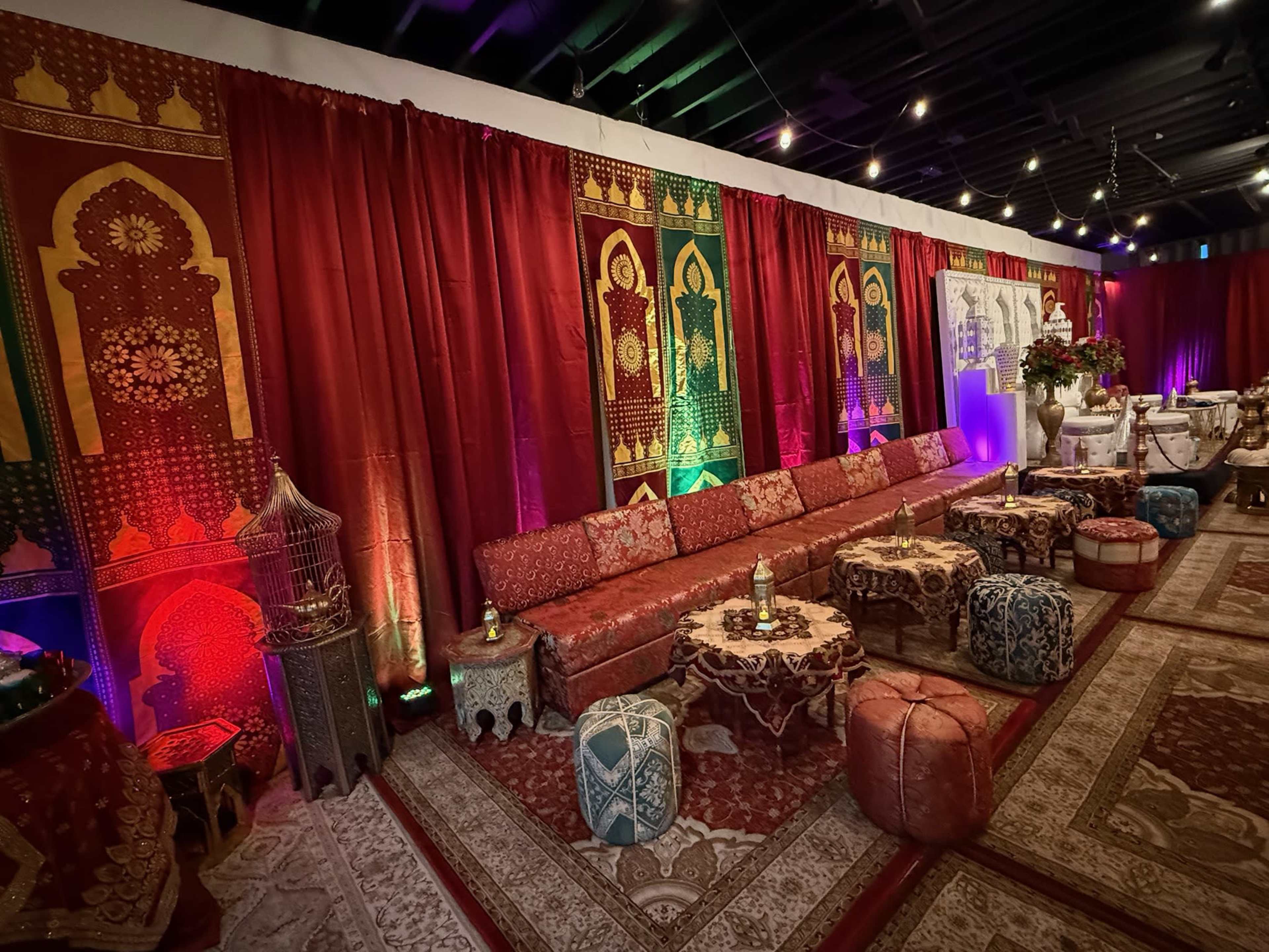 The image shows a decoratively arranged event space featuring rich red fabric drapes, patterned cushions, ornate low tables, and ambient lighting, creating a themed atmosphere.