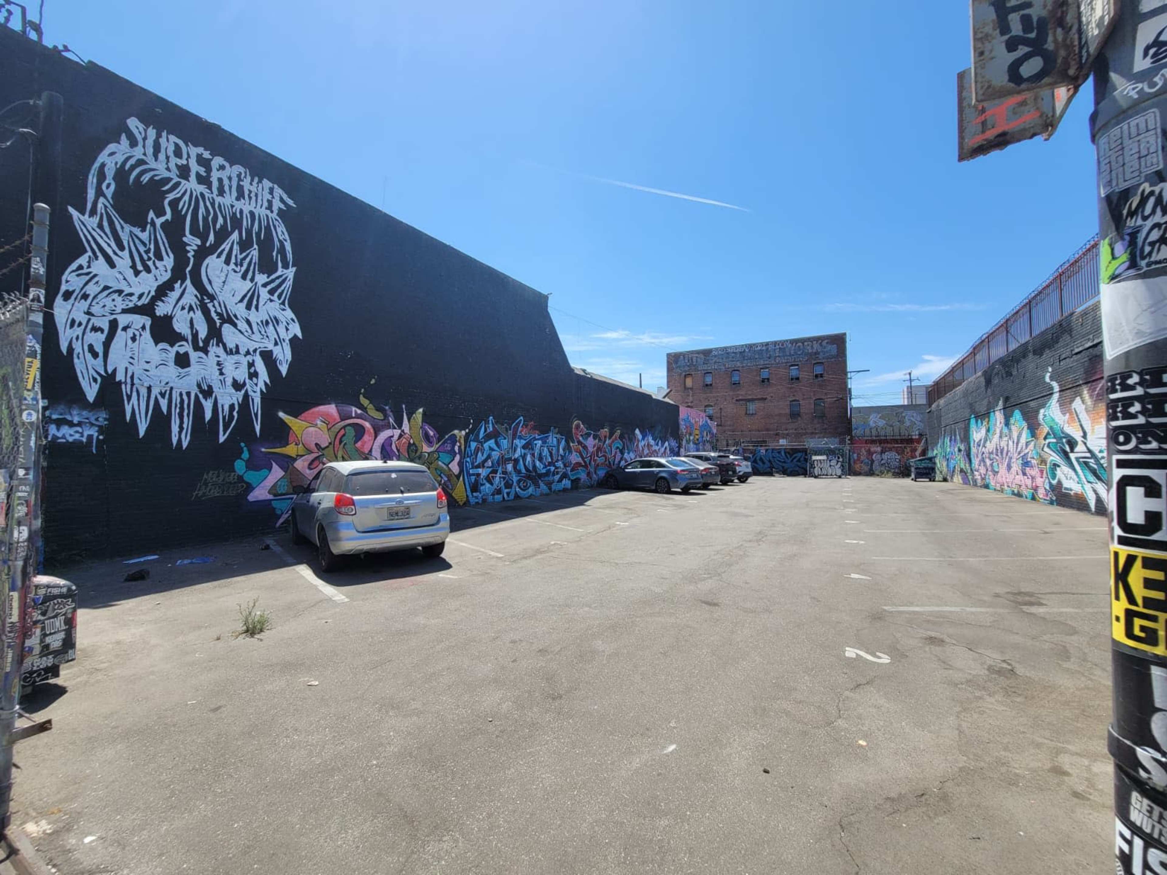 The image shows a graffiti-covered alley with several parked cars and a blue sky in the background.