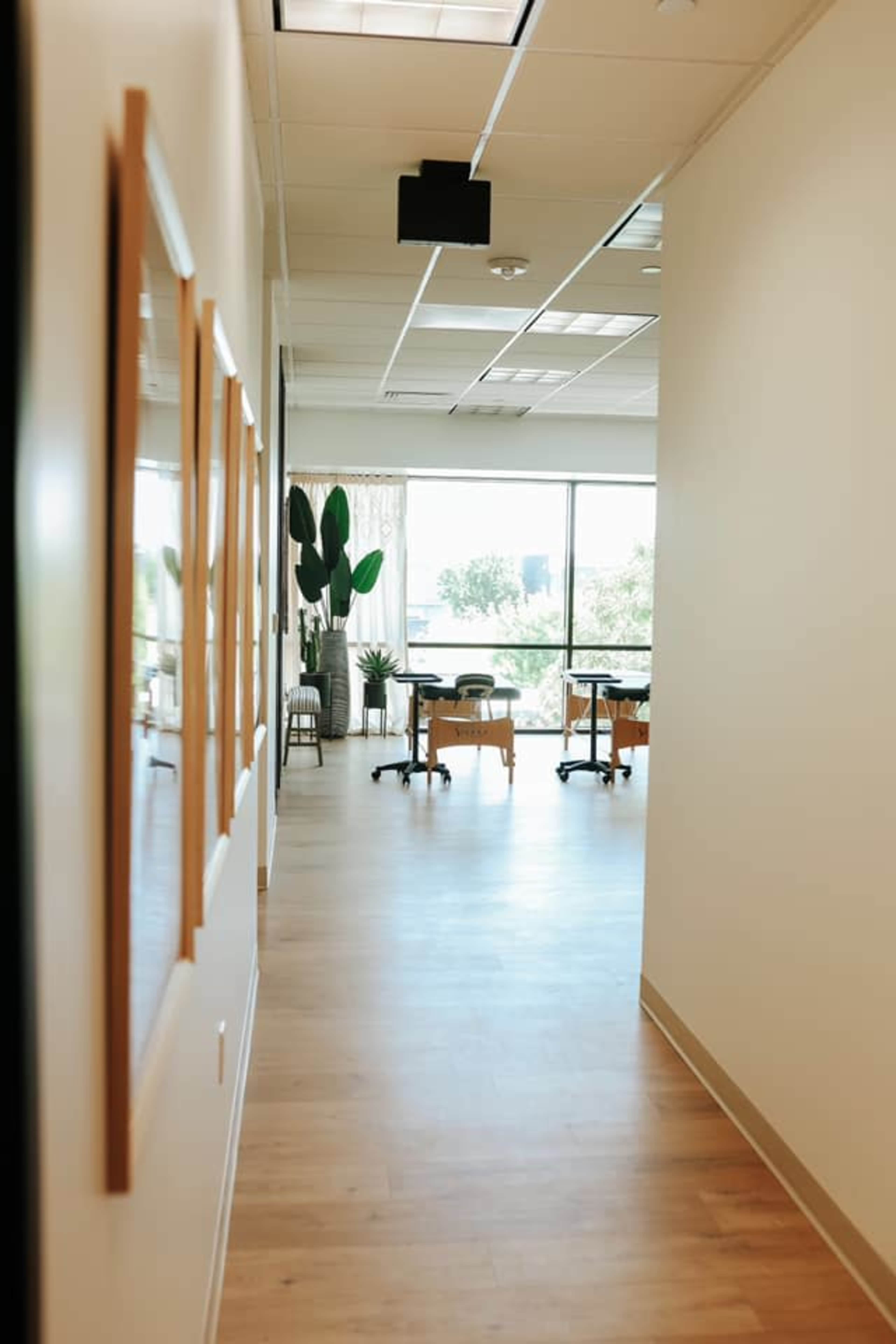 A well-lit hallway leading to a room with tables and plants, framed by large windows.