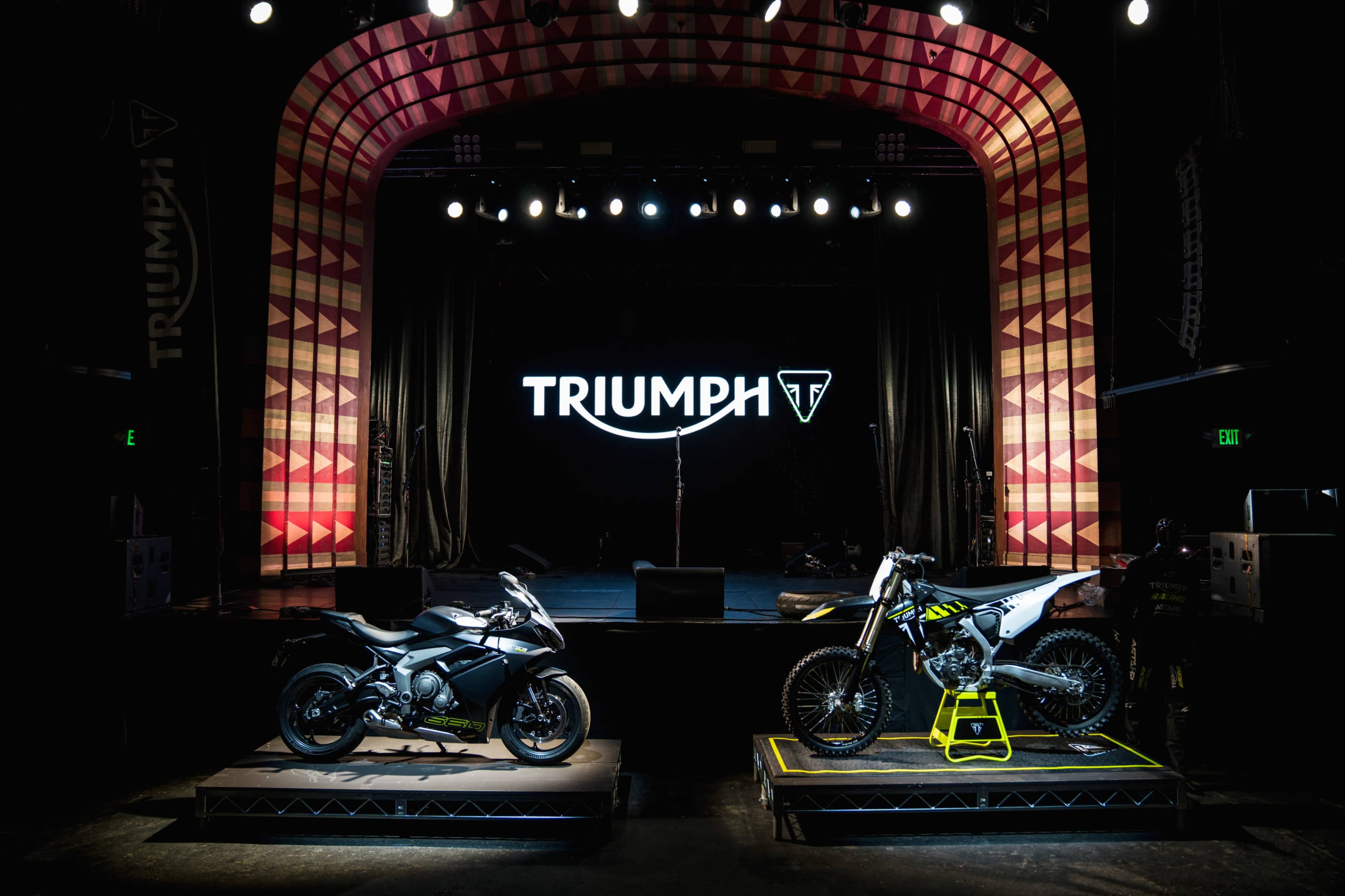 Two Triumph motorcycles displayed on platforms in a dimly lit stage setting, with a large Triumph logo illuminated in the background.