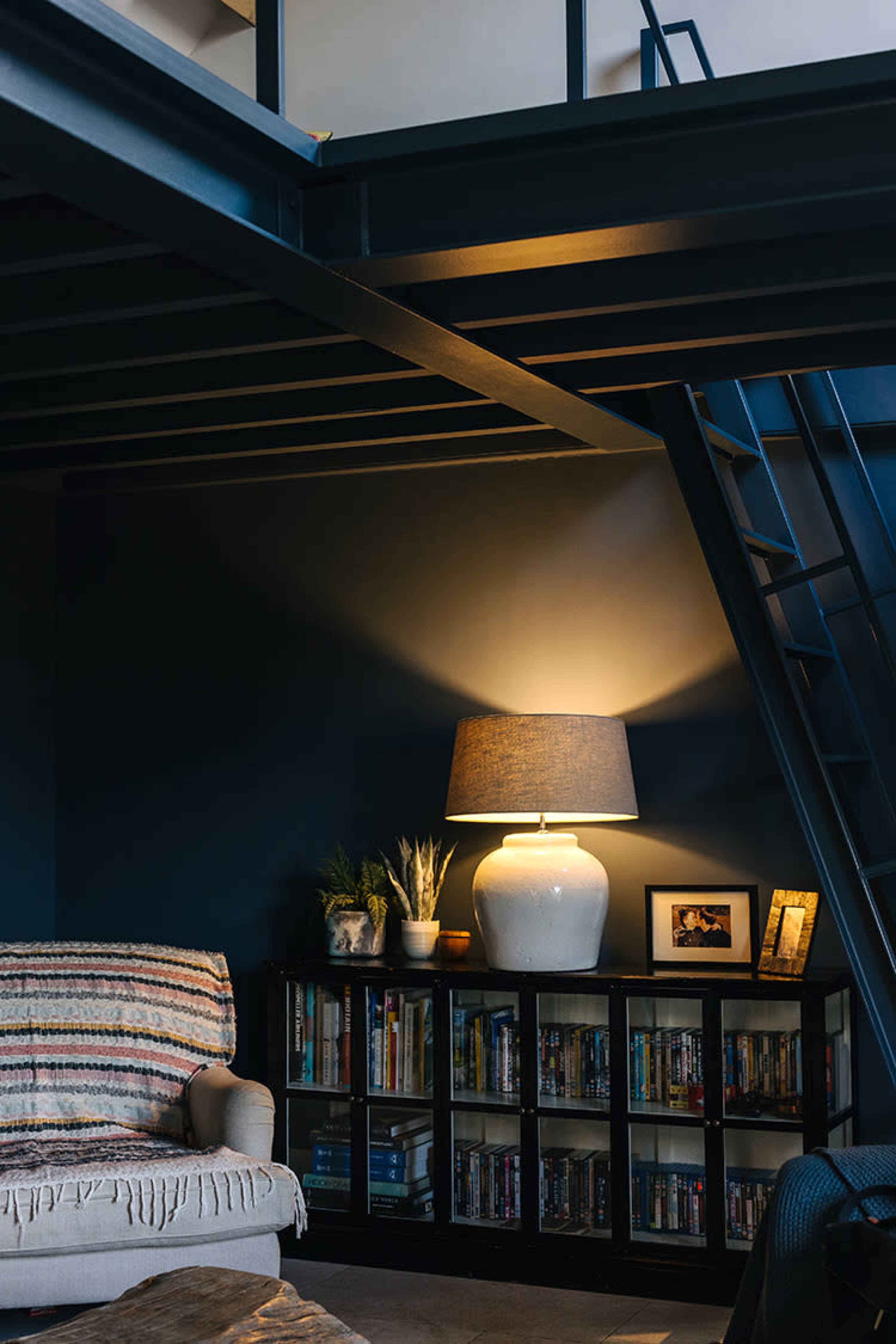A cozy living area features a sofa, a lamp with a lampshade, and a bookshelf filled with books and DVDs, all beneath a staircase.