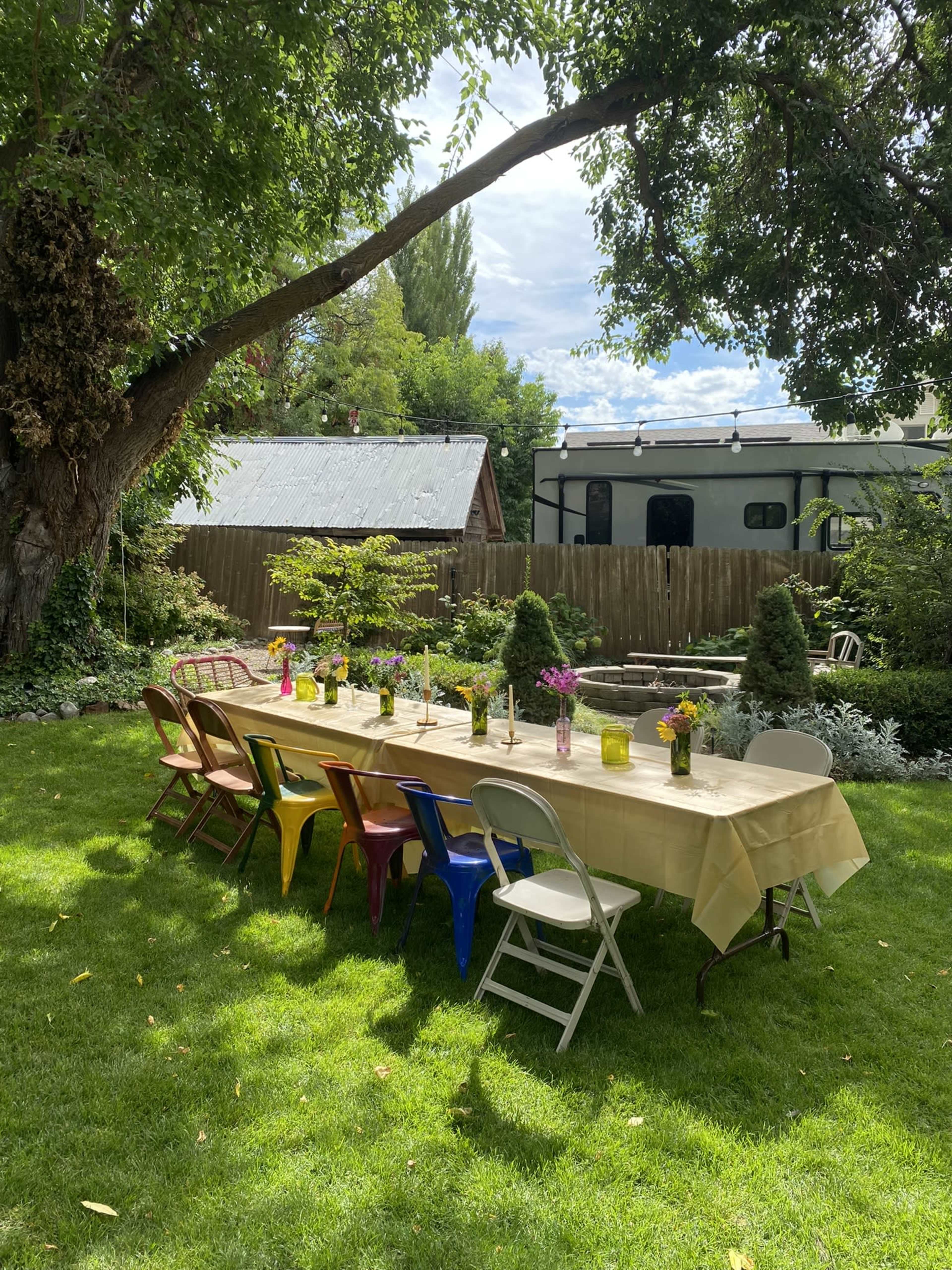 A long table is set with colorful chairs on a green lawn surrounded by trees and plants, with a backdrop of a house and trailer.