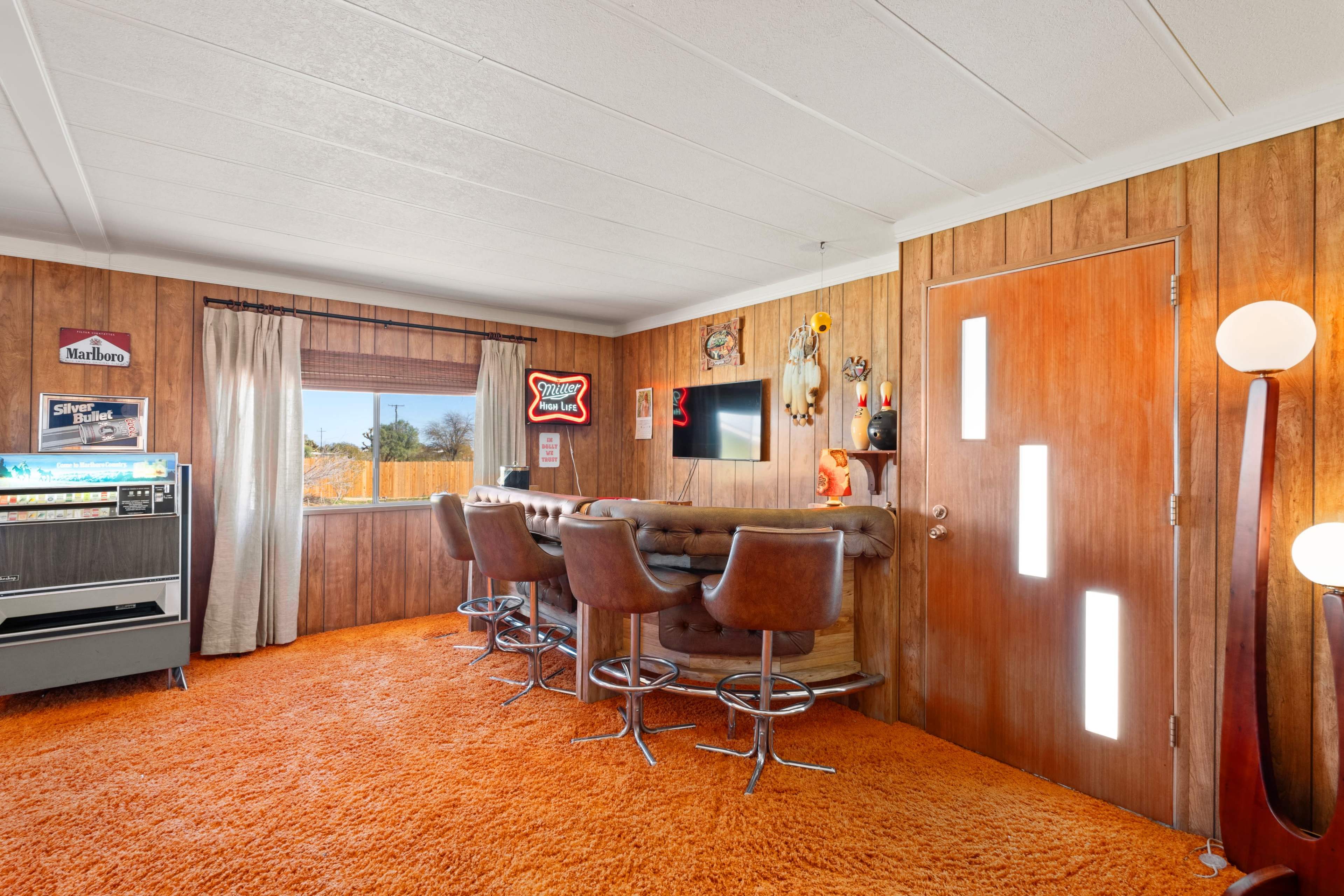 A retro-themed room featuring a bar area with brown leather stools, wood paneling, an orange carpet, and a vintage jukebox.