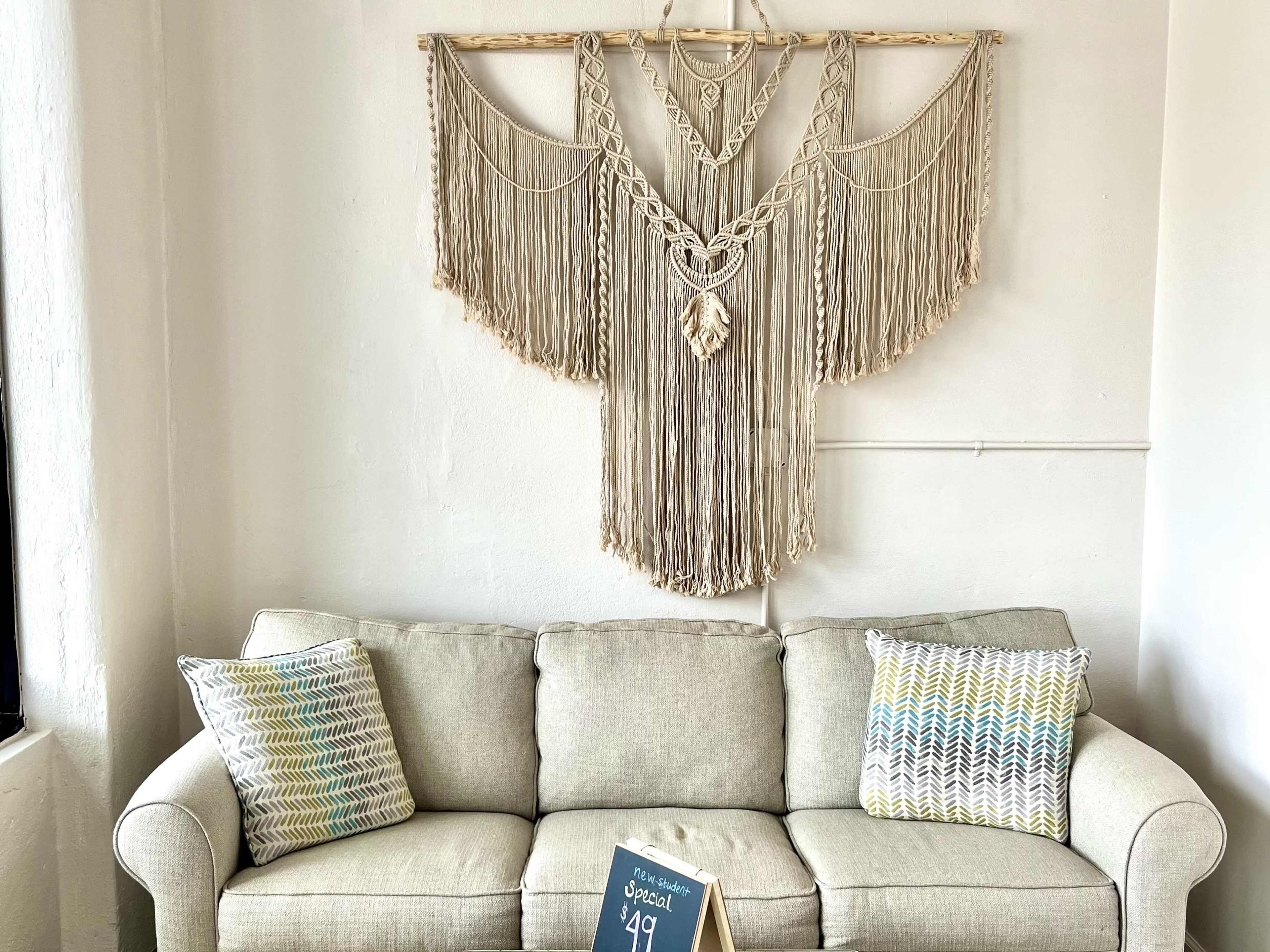A macramé wall hanging is displayed above a gray sofa, which is flanked by two decorative pillows and a small sign on the floor.