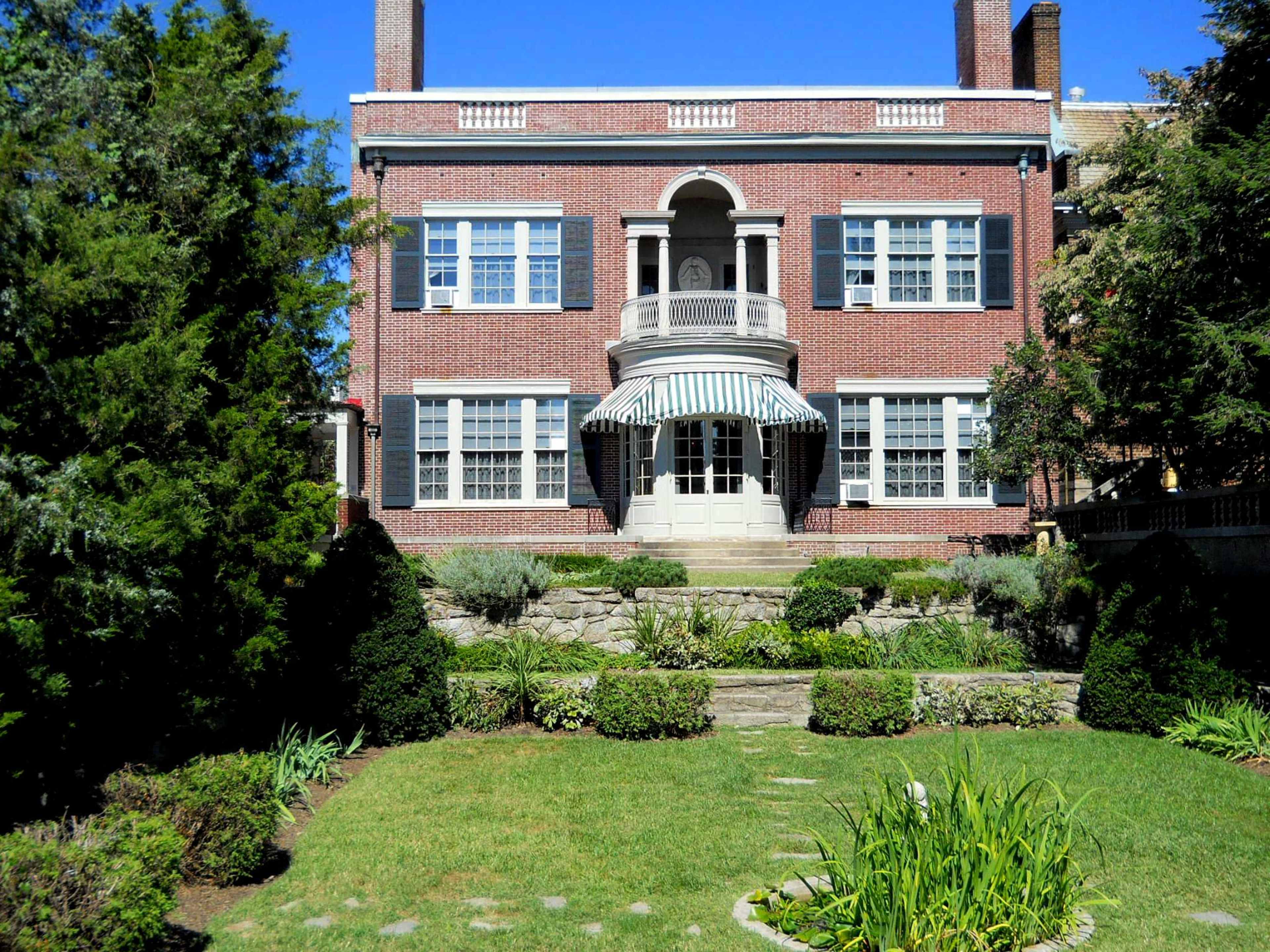 A brick house with a covered porch, surrounded by landscaped gardens and pathways.