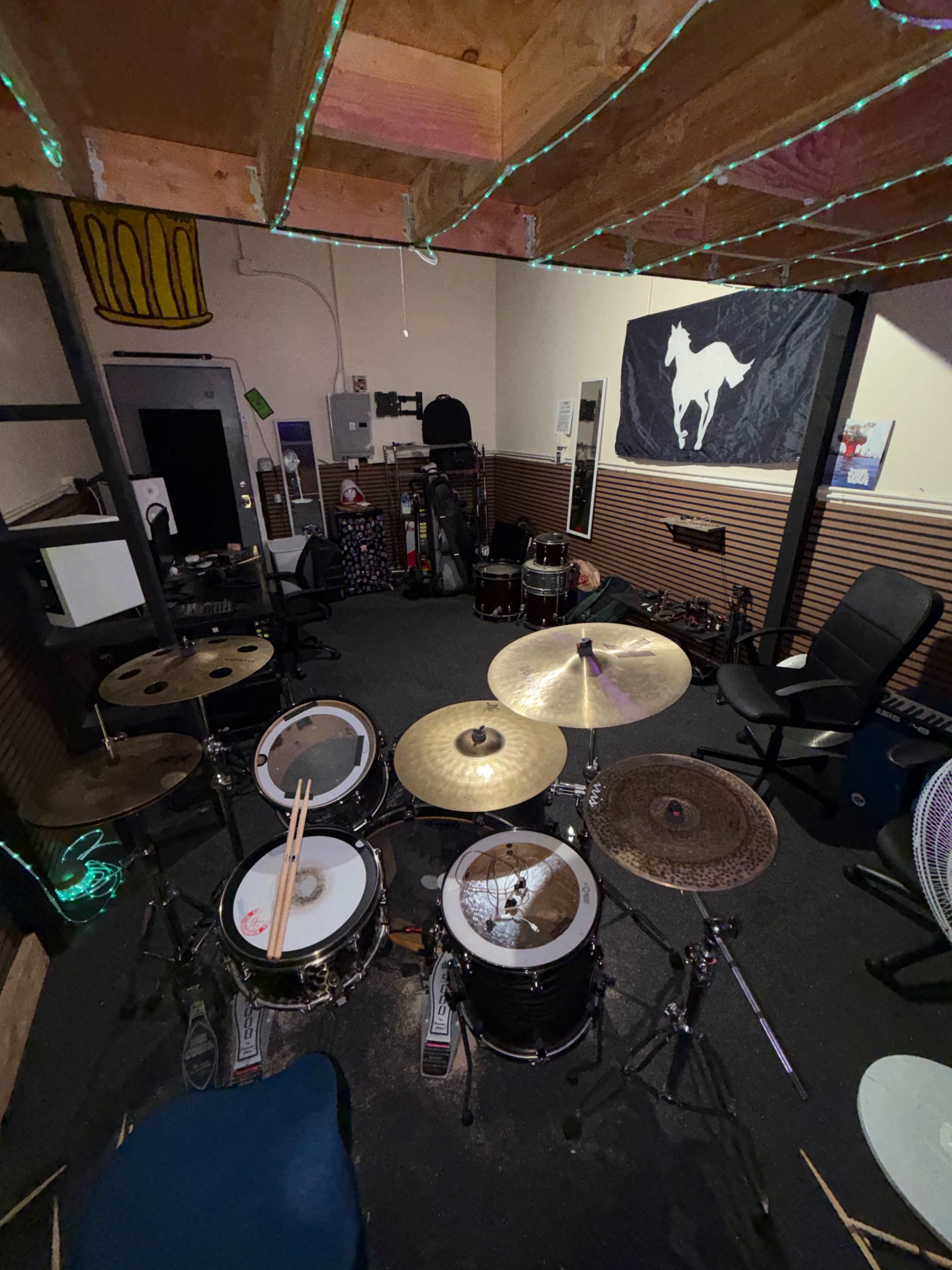 The image shows a home recording studio with a drum set in the foreground and various music equipment and chairs arranged around the room.