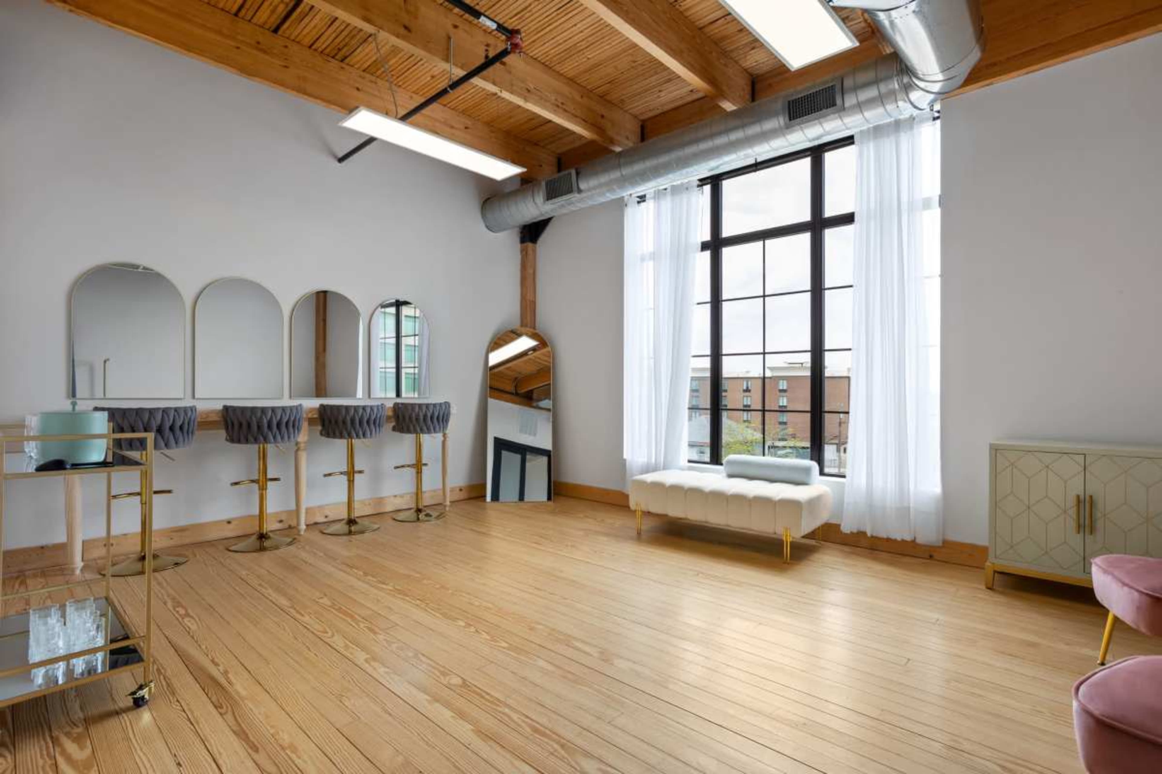 The image shows a spacious room with hardwood flooring, large windows, and contemporary furnishings, including a seating area and dressing mirrors.