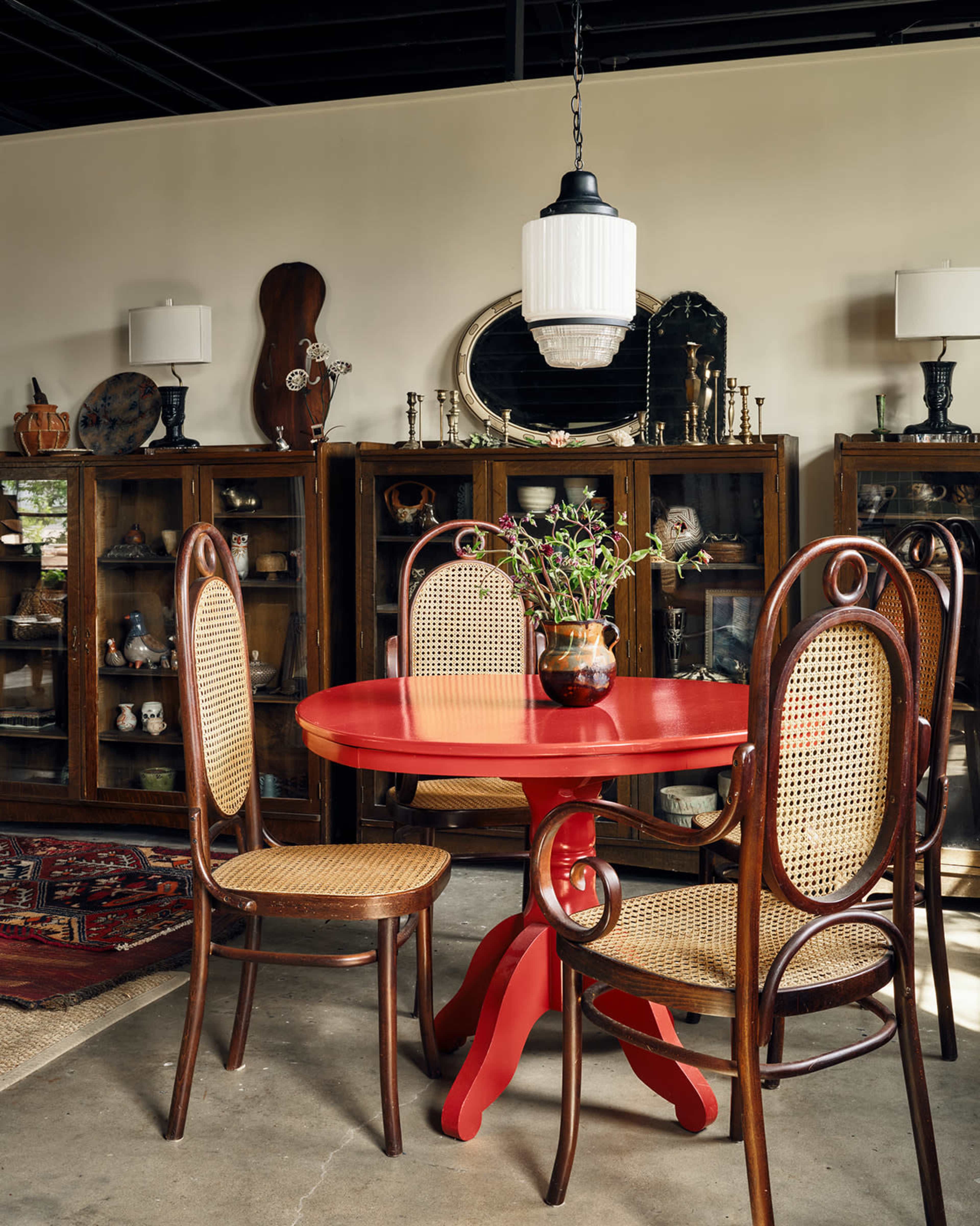 A red table with two caned chairs and a vase of flowers is set in a room featuring wooden display cabinets and antique decor.
