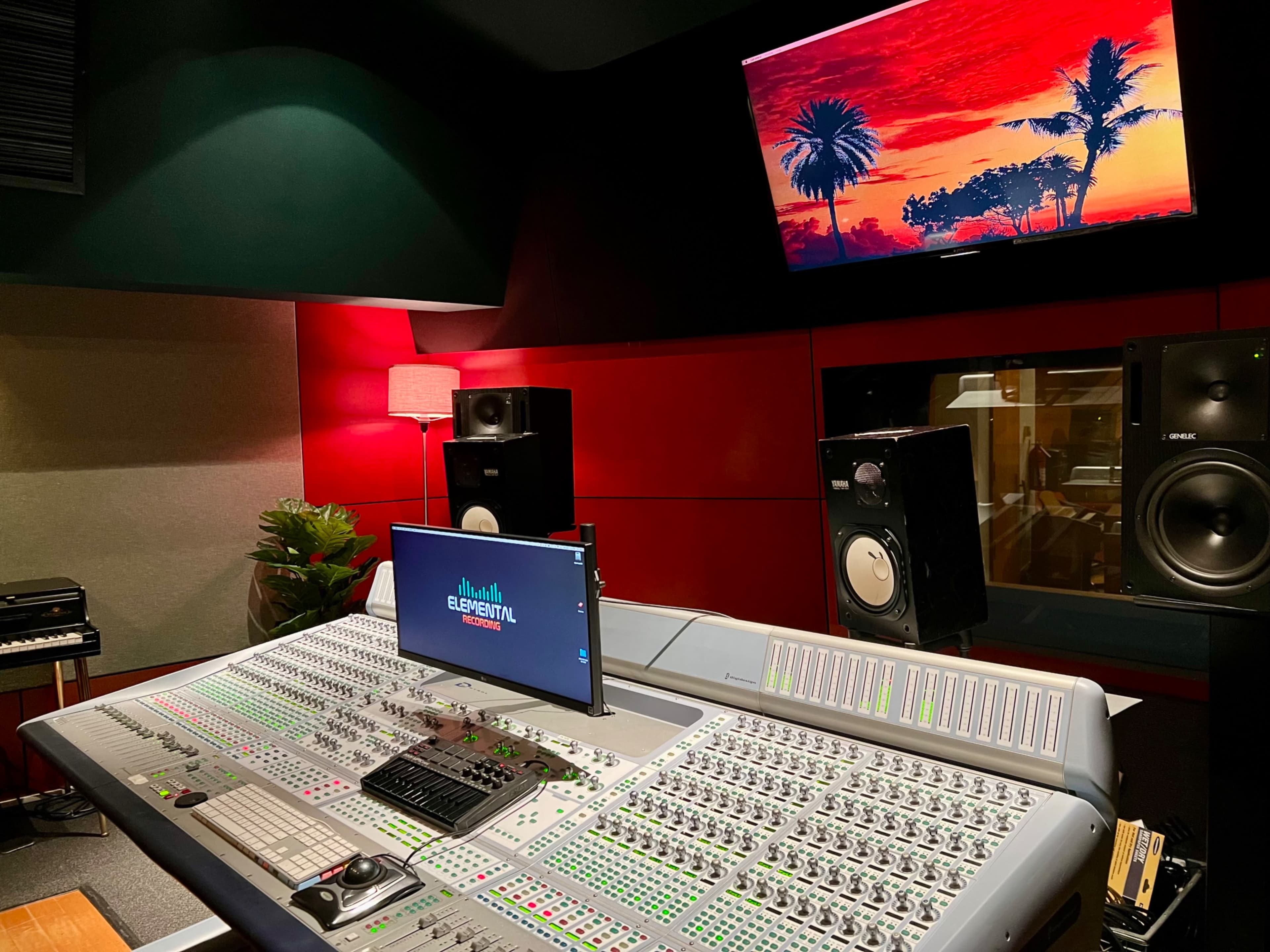 The image shows a music studio with a large mixing console, two studio monitors, a television displaying a sunset scene, and ambient lighting.