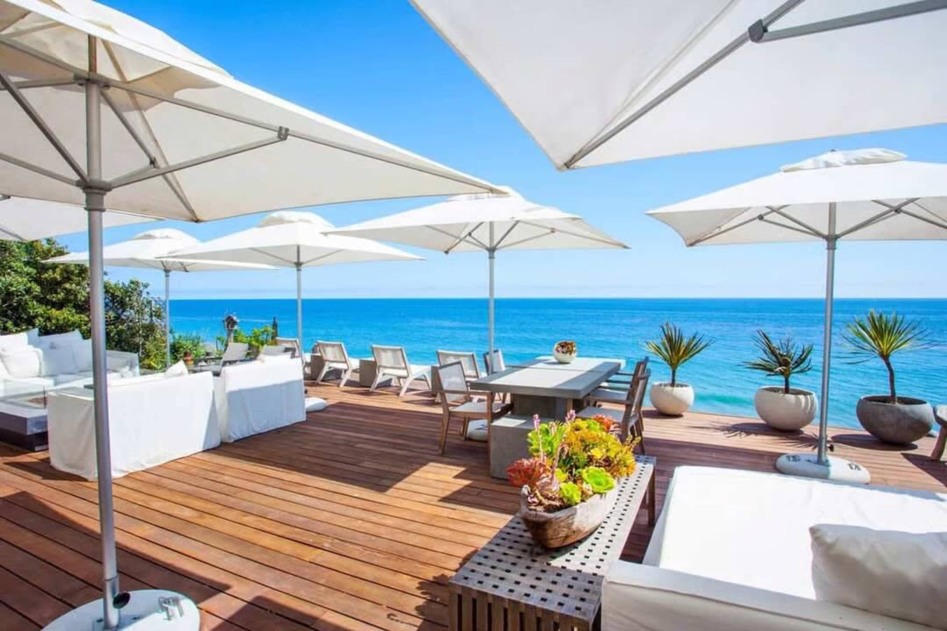A spacious wooden deck features lounge chairs and dining areas, shaded by white umbrellas, overlooking a serene ocean view.