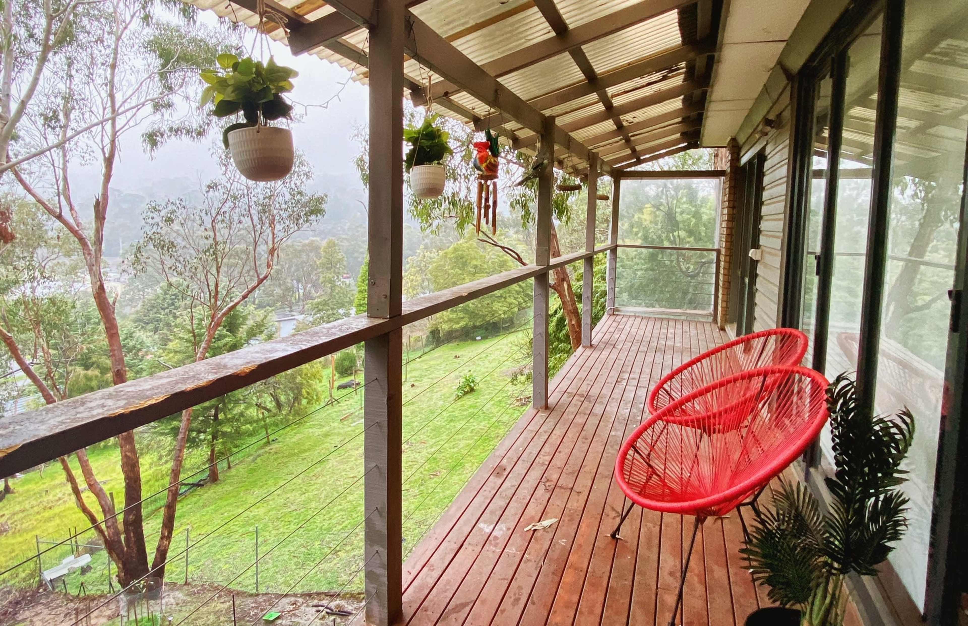 The image shows a wooden porch with two red hanging chairs, surrounded by potted plants and a view of a green landscape beyond.