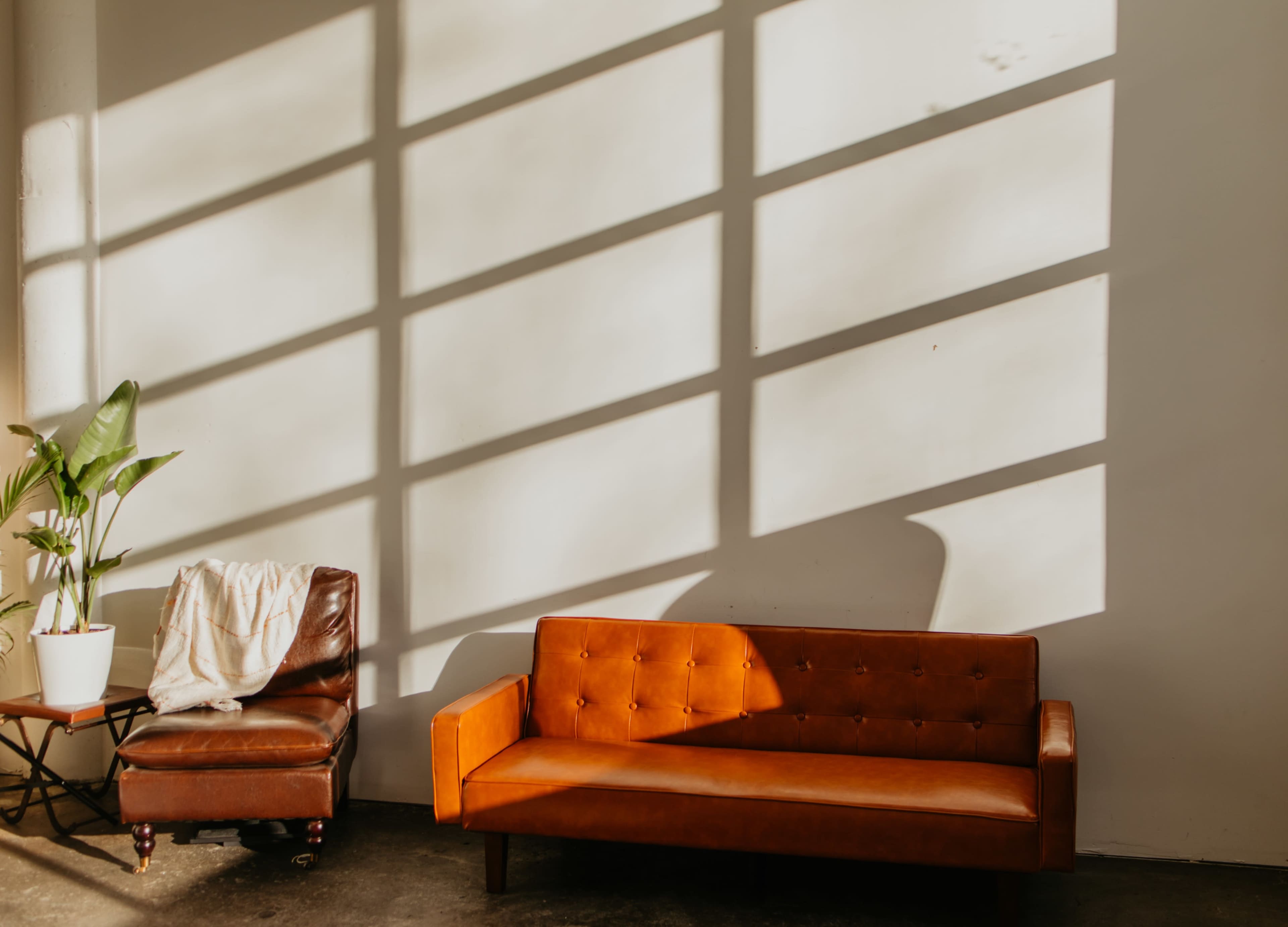 A brown leather armchair and an orange sofa are positioned against a wall, with geometric shadows cast by sunlight filtering through a window.
