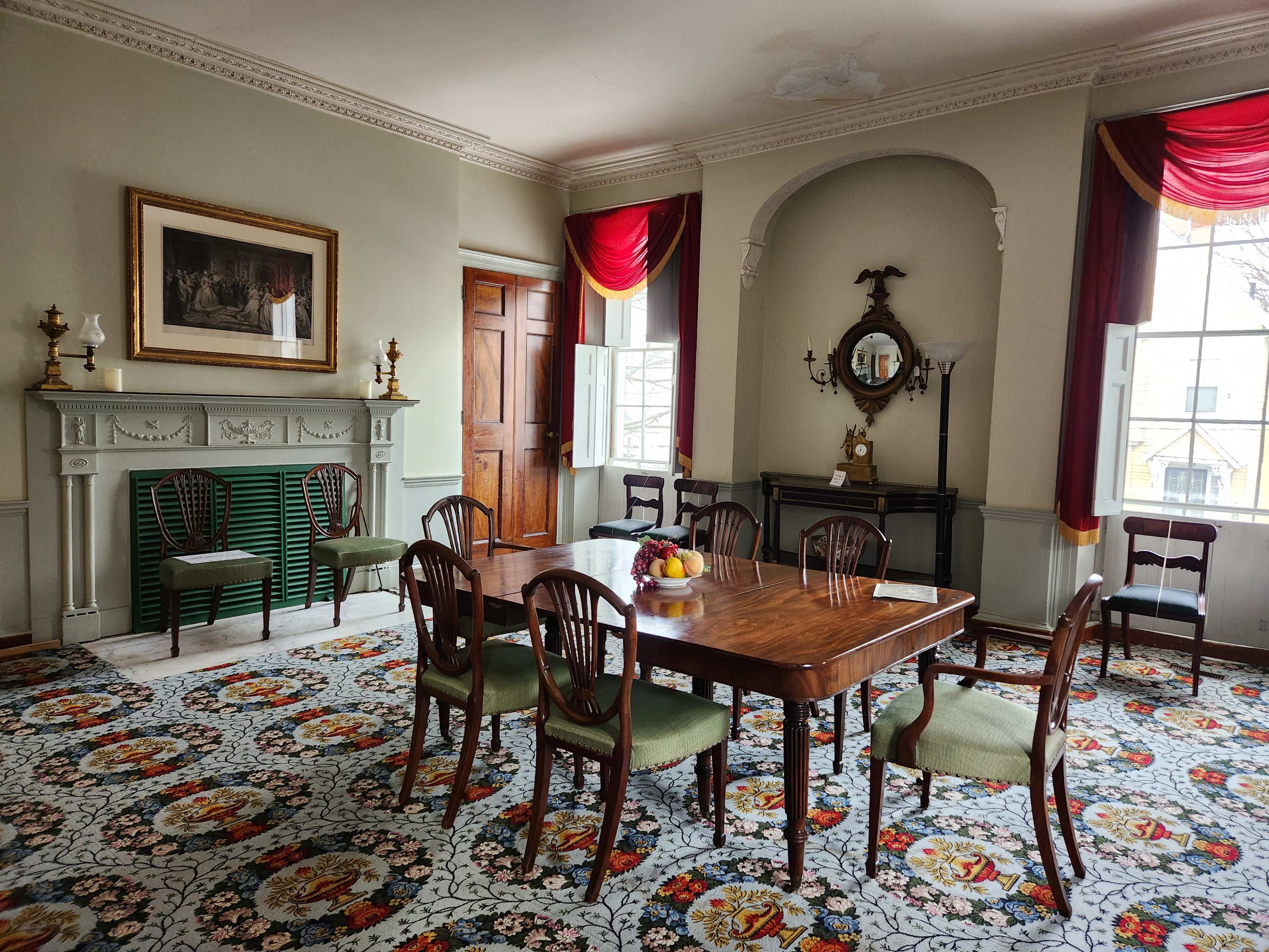 The dining room features a wooden table surrounded by eight chairs, with a decorative rug covering the floor and large windows dressed with red drapes.
