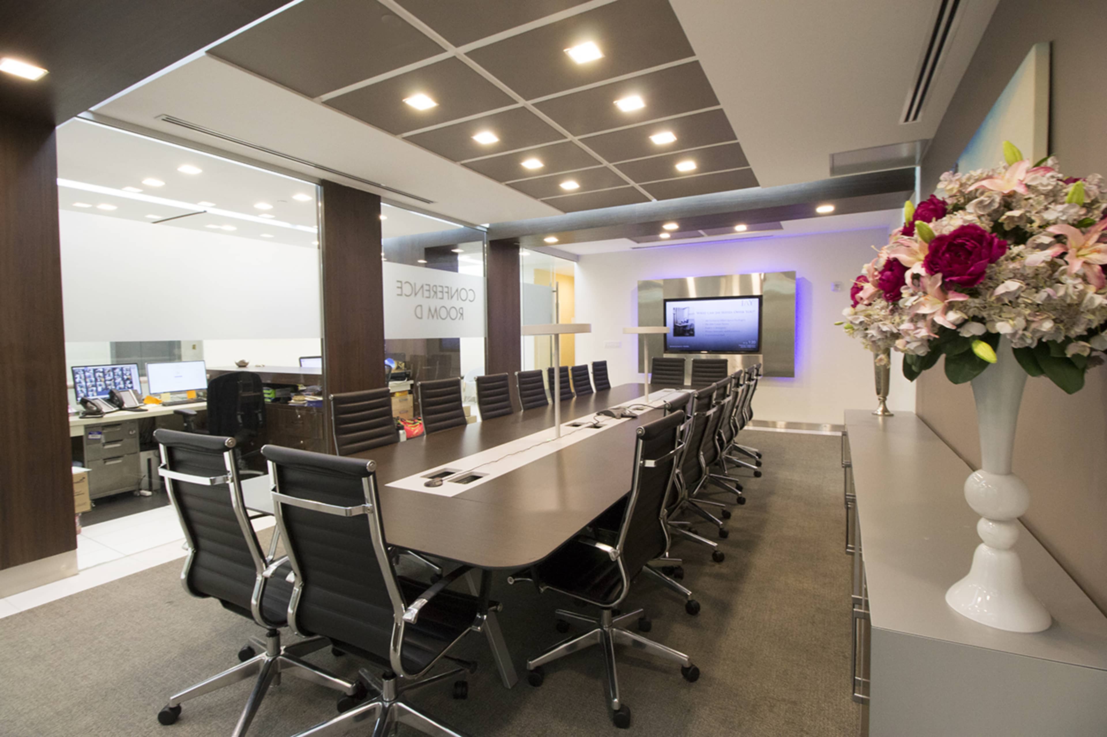 A large conference room features a long table surrounded by black chairs, with a wall-mounted screen and computers visible in the background.