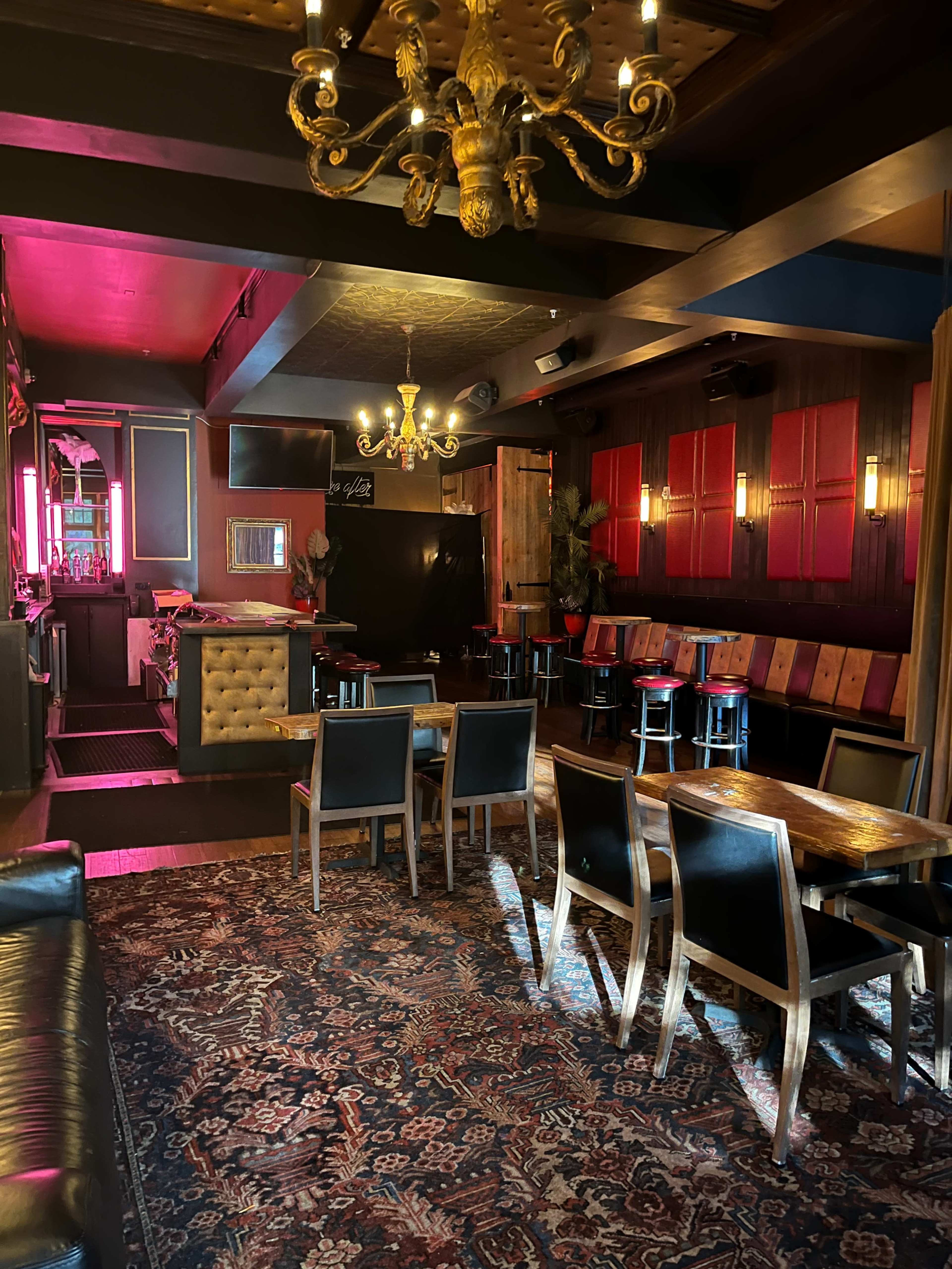 The image shows the interior of a dimly lit bar featuring wooden furnishings, ornate chandeliers, and a decorative rug on the floor.