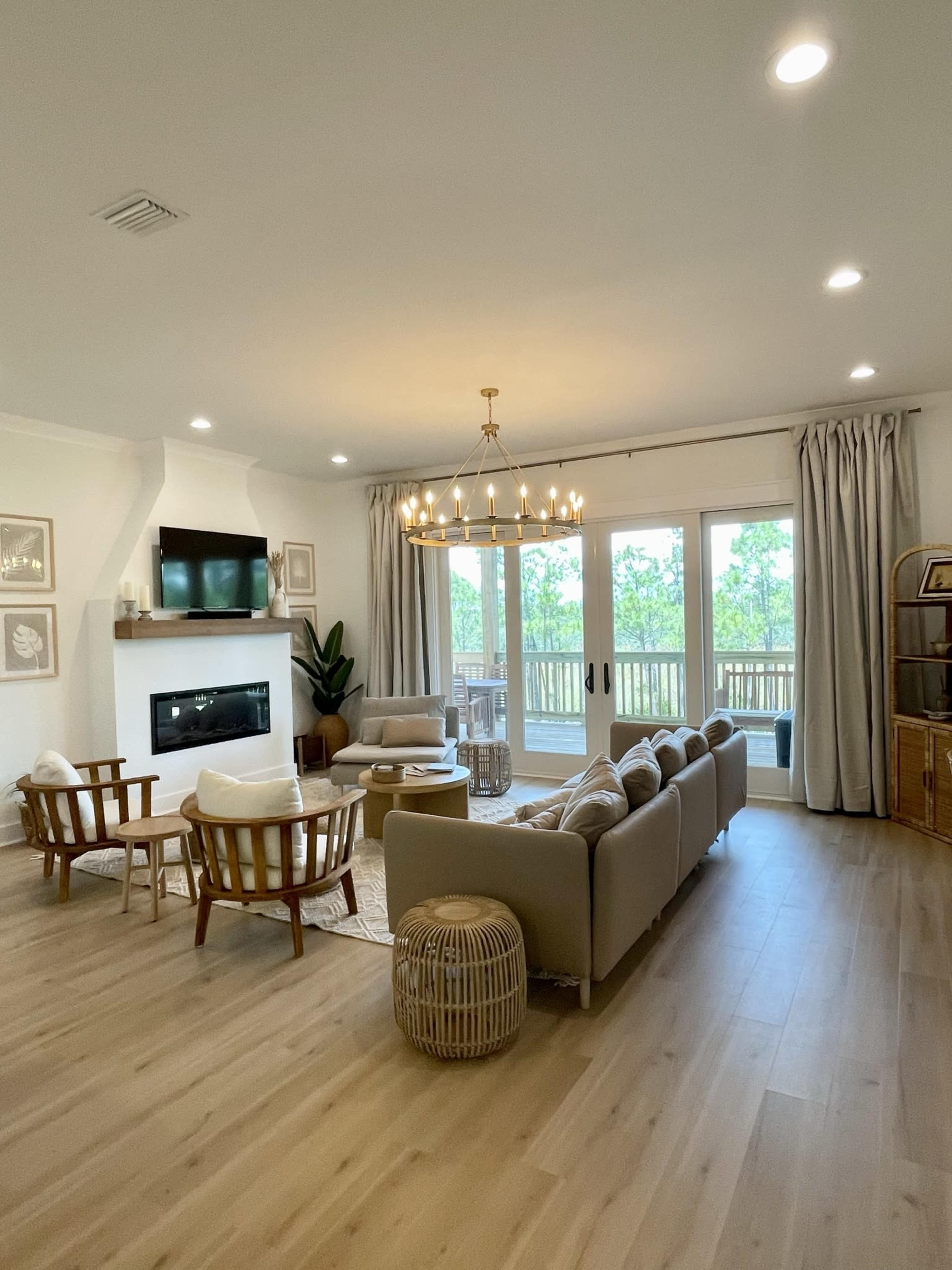 The living room features a sectional sofa, a coffee table, and a large light fixture, with large windows showcasing a view of greenery outside.