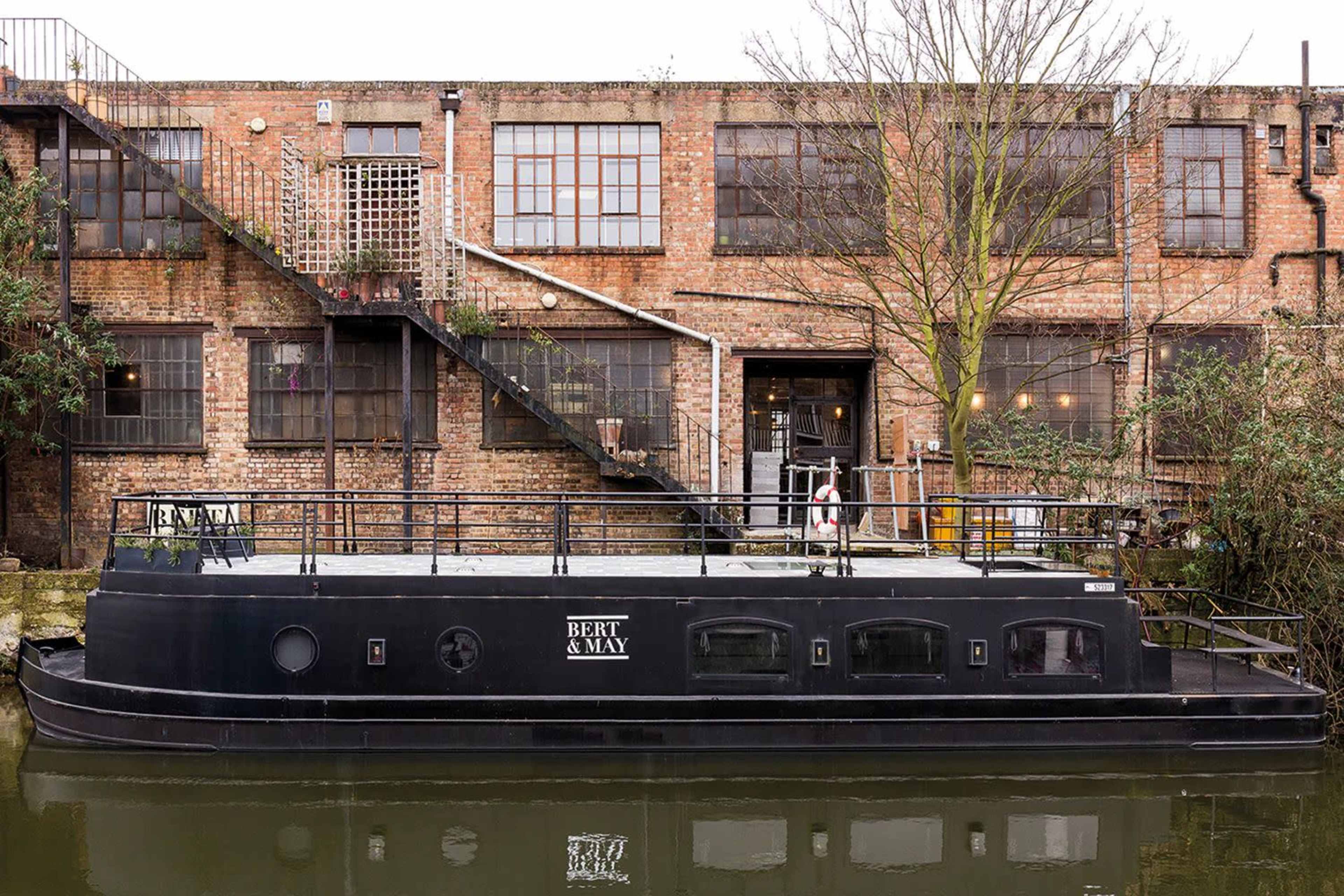 A black barge named "BERT & MAY" is moored beside a brick warehouse with large windows and an outdoor staircase.