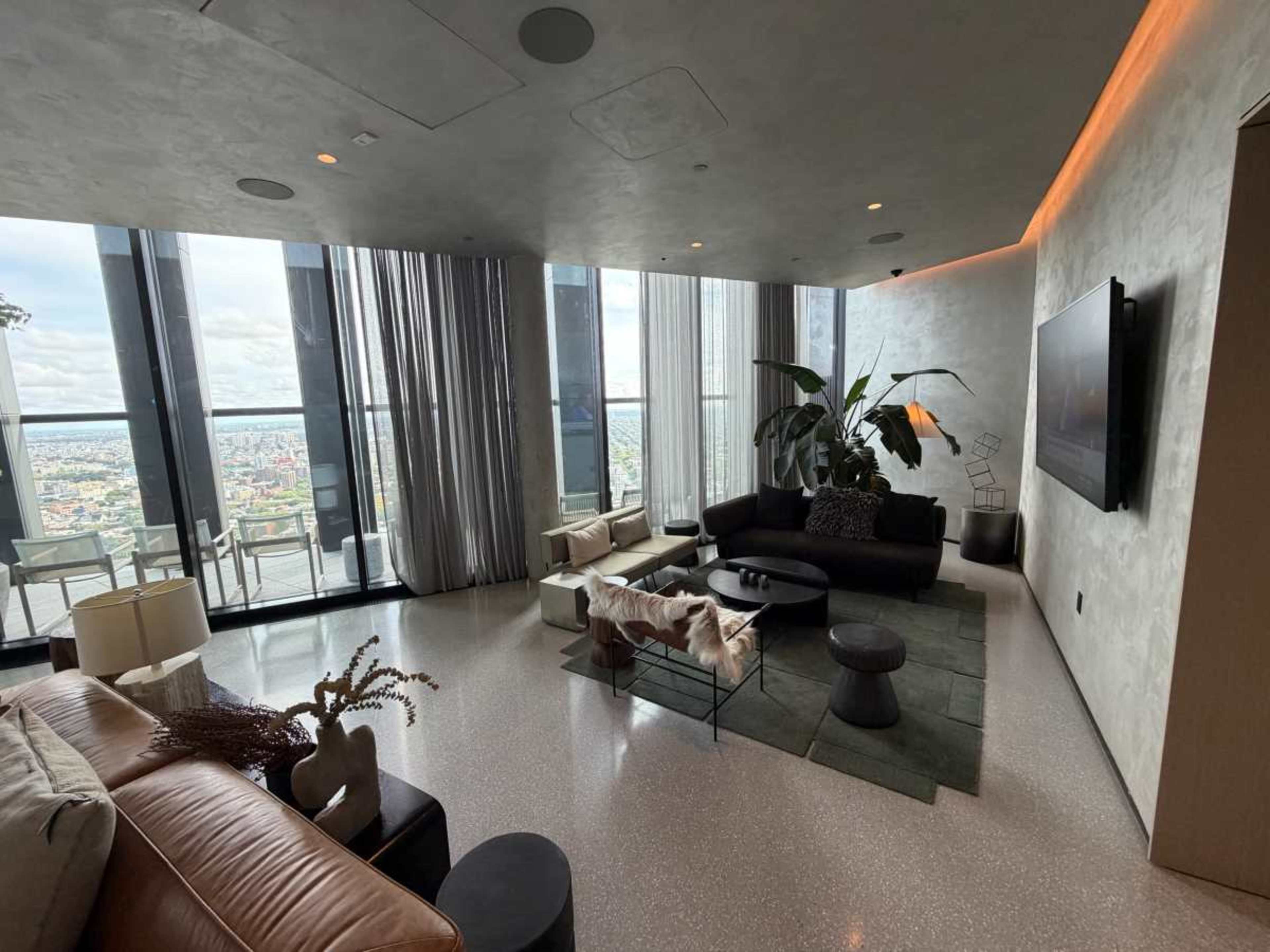 The image shows a modern living room with large windows offering a city view, featuring a mix of comfortable seating, a coffee table, and decorative plants.