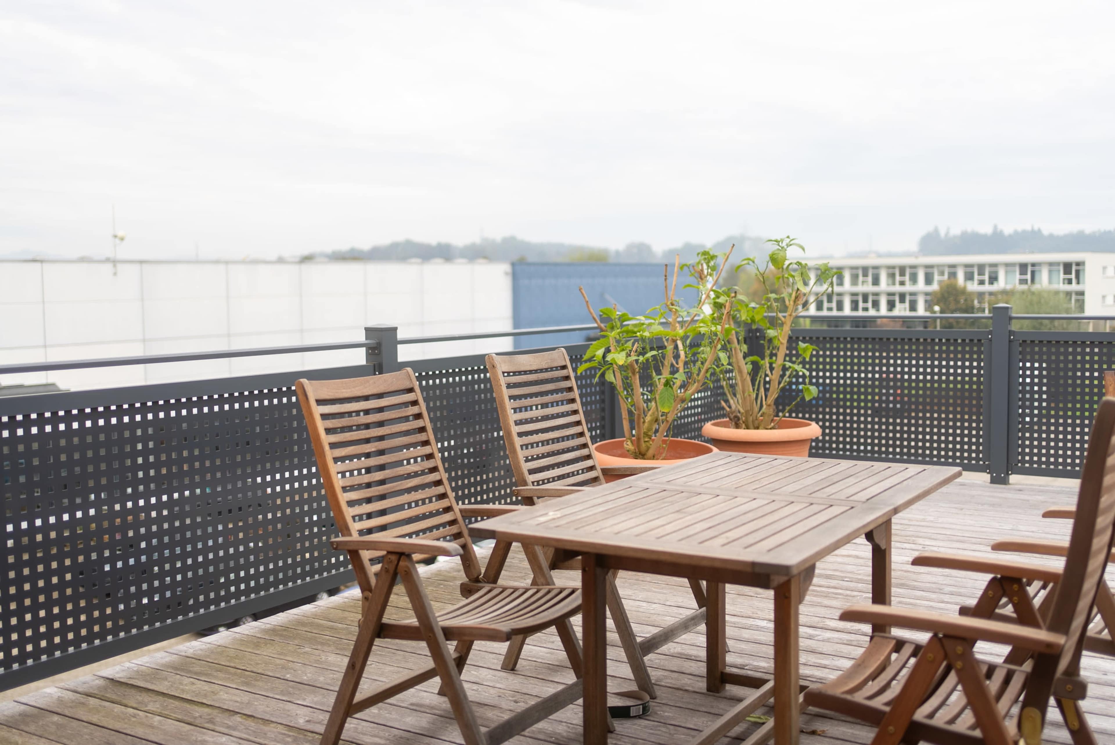 A wooden patio set with a table and chairs is positioned on a deck, overlooking potted plants and a distant urban landscape.
