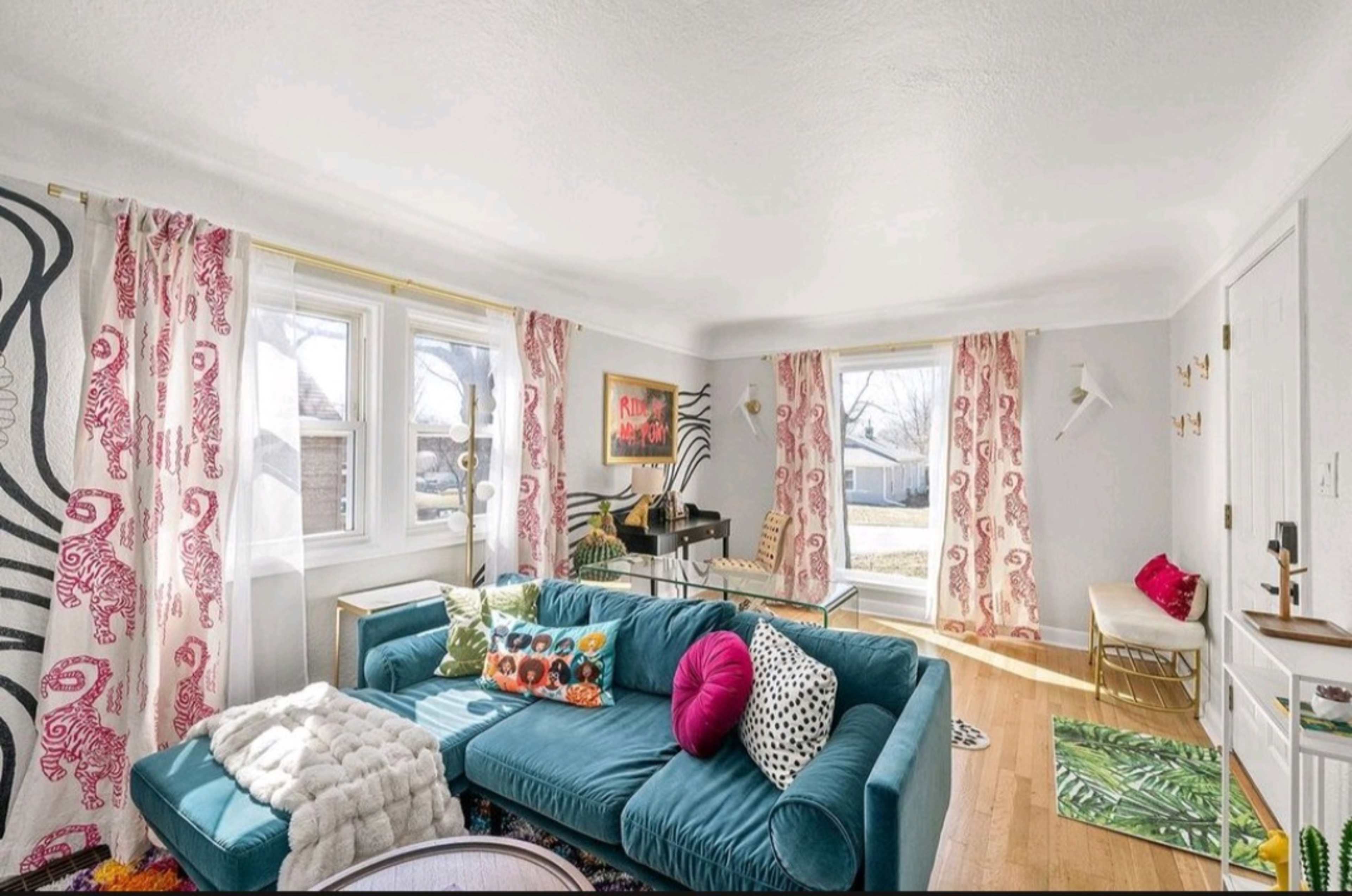 The living room features a turquoise sofa, a glass coffee table, colorful cushions, and large windows with patterned curtains.