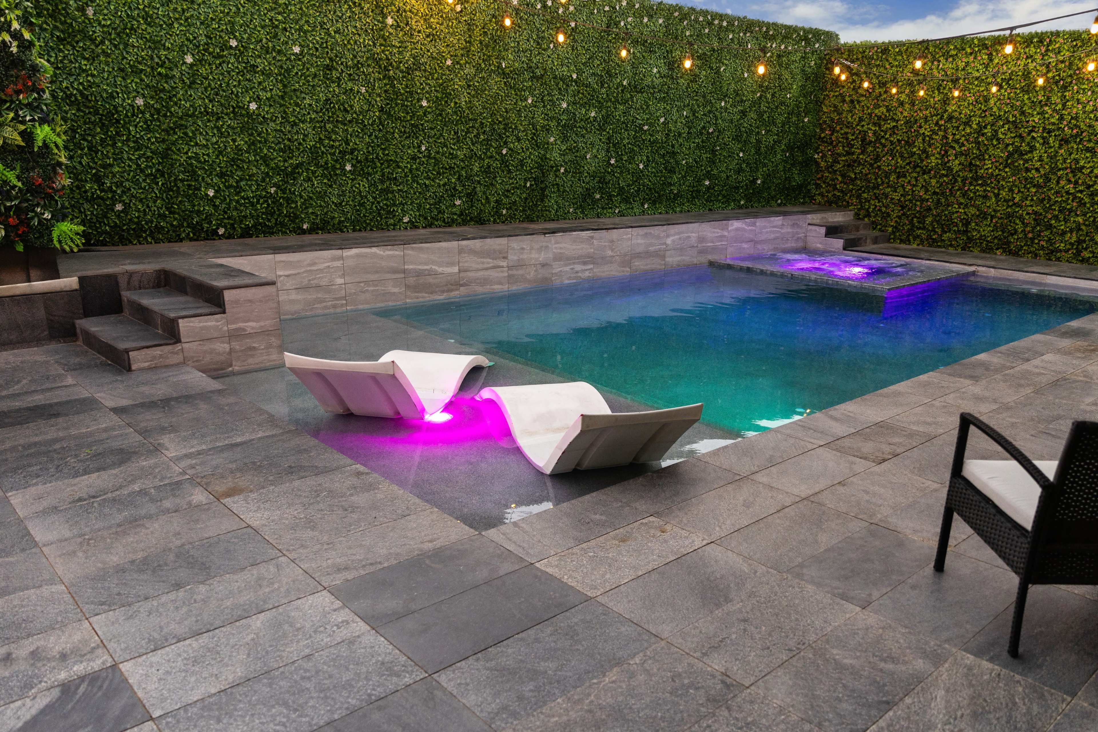 The image shows a modern swimming pool surrounded by stone tiles, with two white lounge chairs partially submerged and purple LED lights illuminating the water.