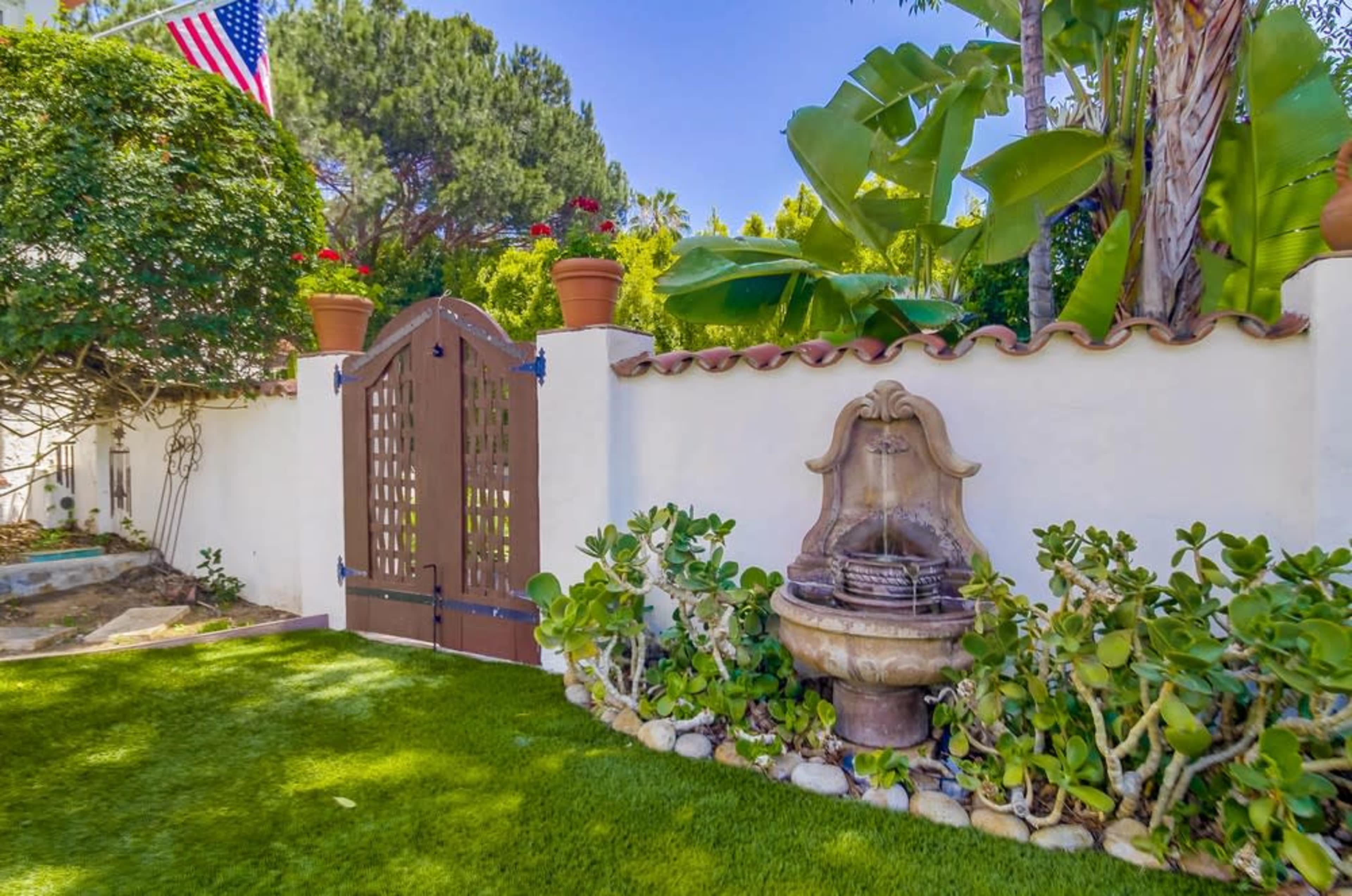 A gated entrance with a wooden door is set against a backdrop of lush greenery and a stone fountain in a well-maintained garden.