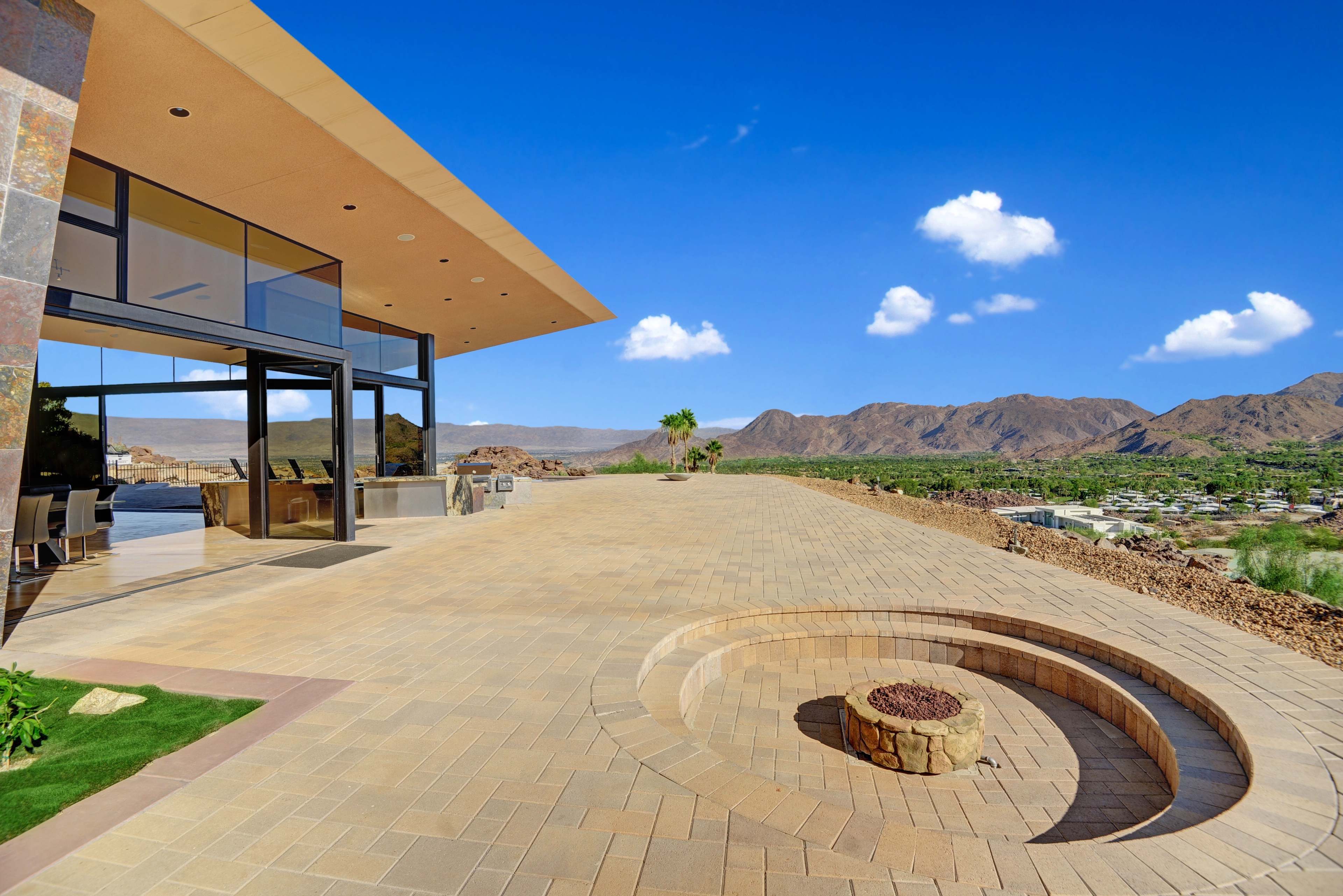 A modern building with large windows overlooks a spacious patio featuring a circular fire pit and a backdrop of mountains.