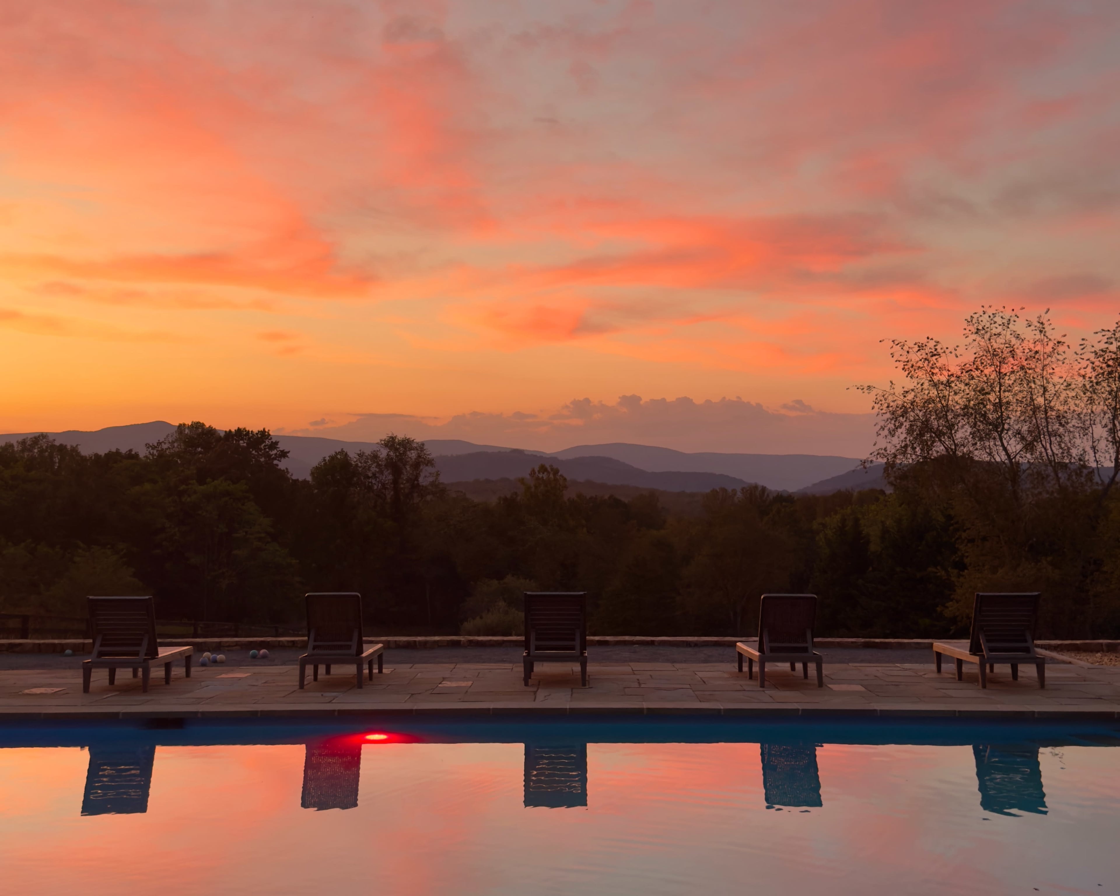 Lounge chairs line the edge of a swimming pool, reflecting the vibrant colors of a sunset over distant mountains.