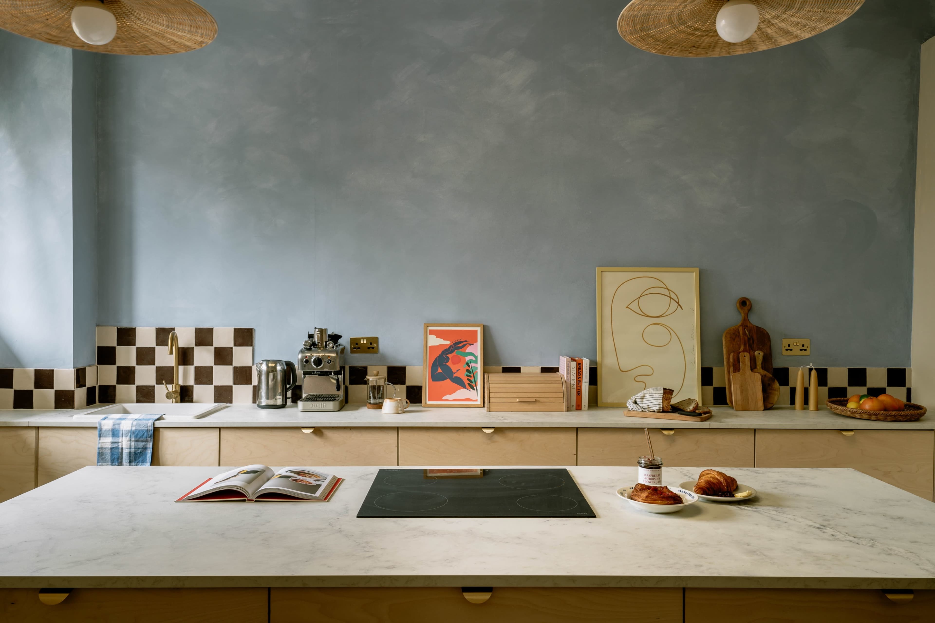 A modern kitchen features a large marble island with a stovetop, an open cookbook, and a plate of croissants, surrounded by minimalist decor and wooden elements.