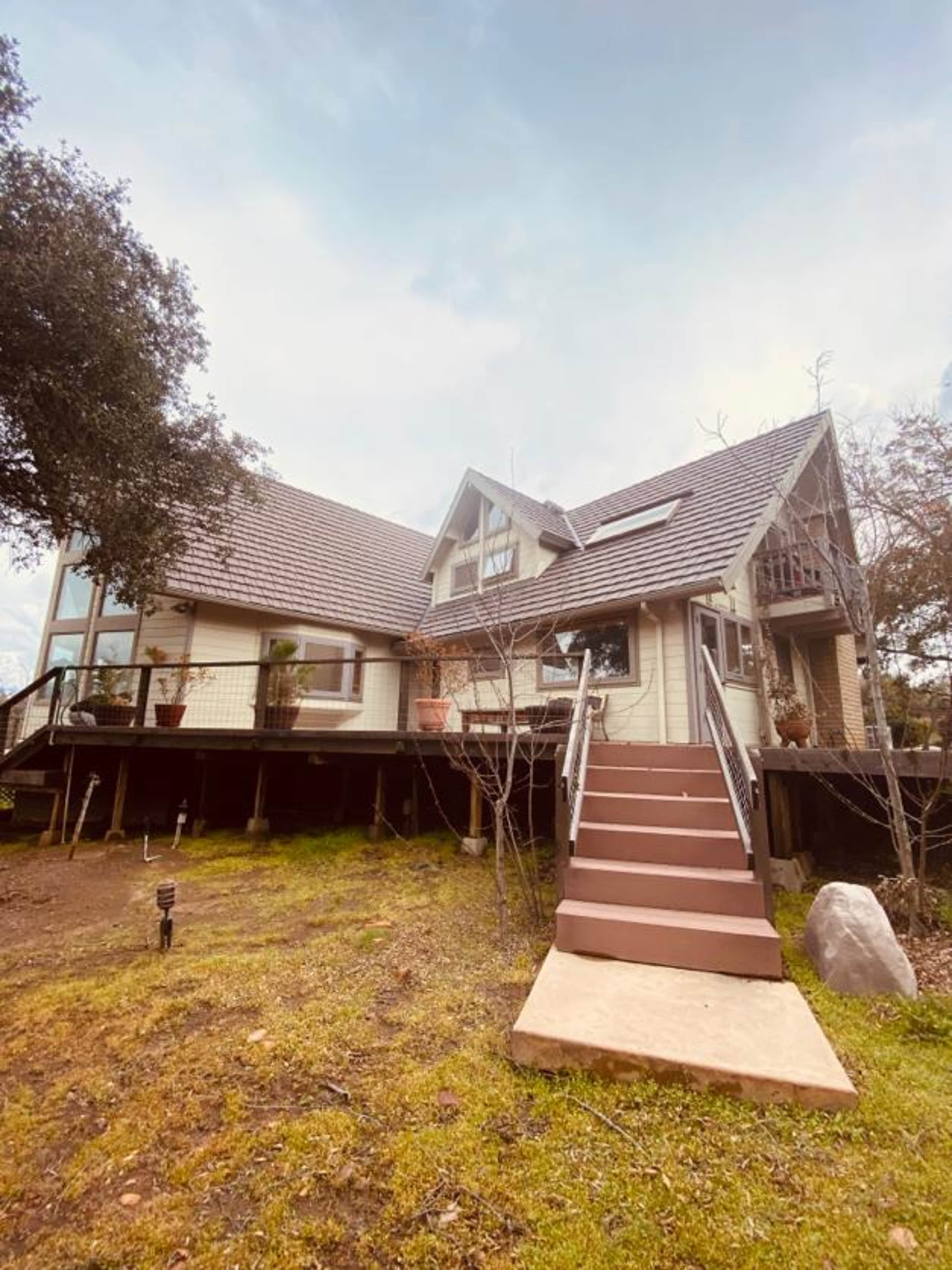 A two-story house with a steep roof and a front staircase is surrounded by grass and trees.