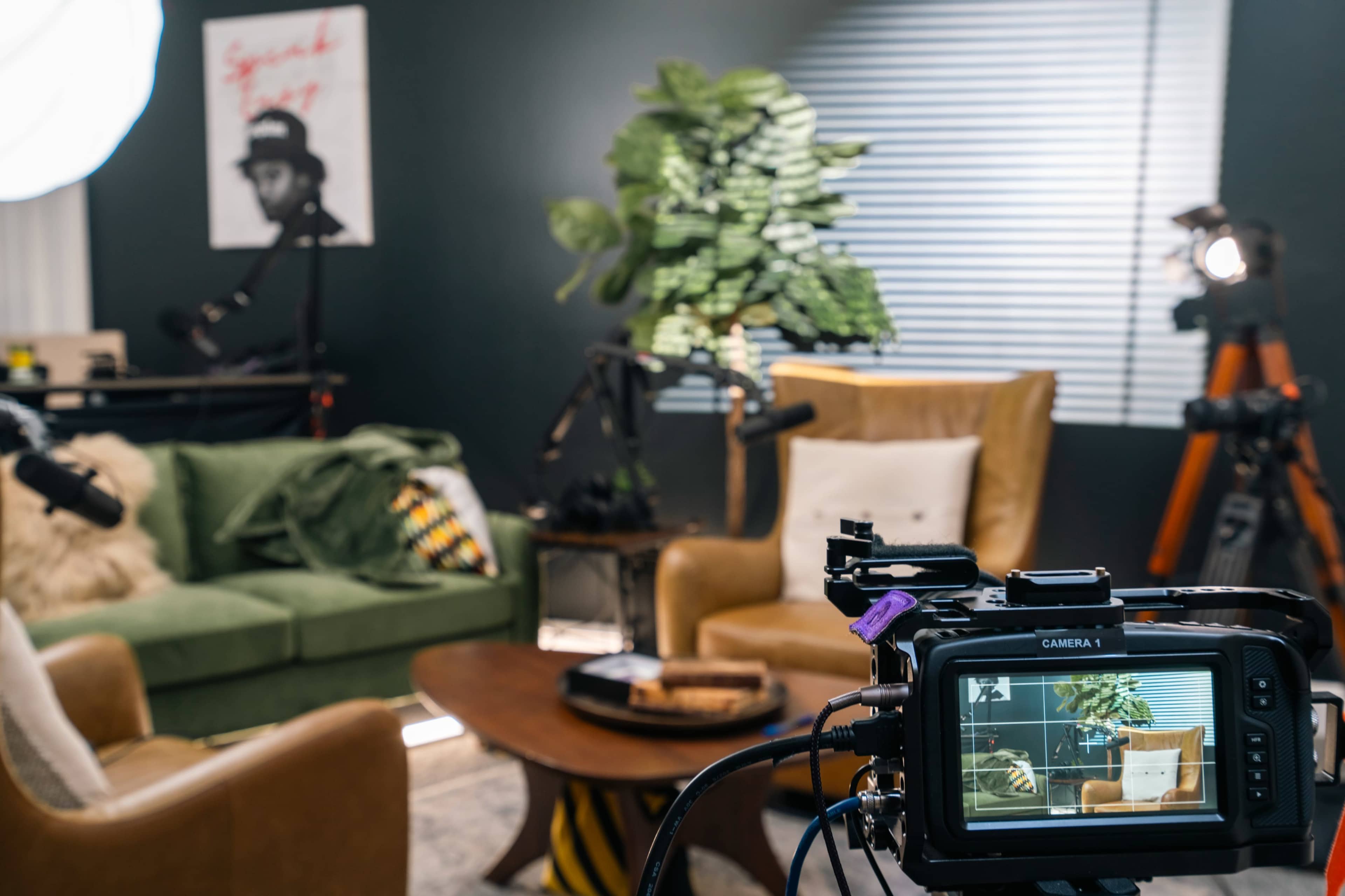 A well-lit recording setup featuring a camera aimed at a cozy living area with two chairs, a coffee table, and indoor plants.