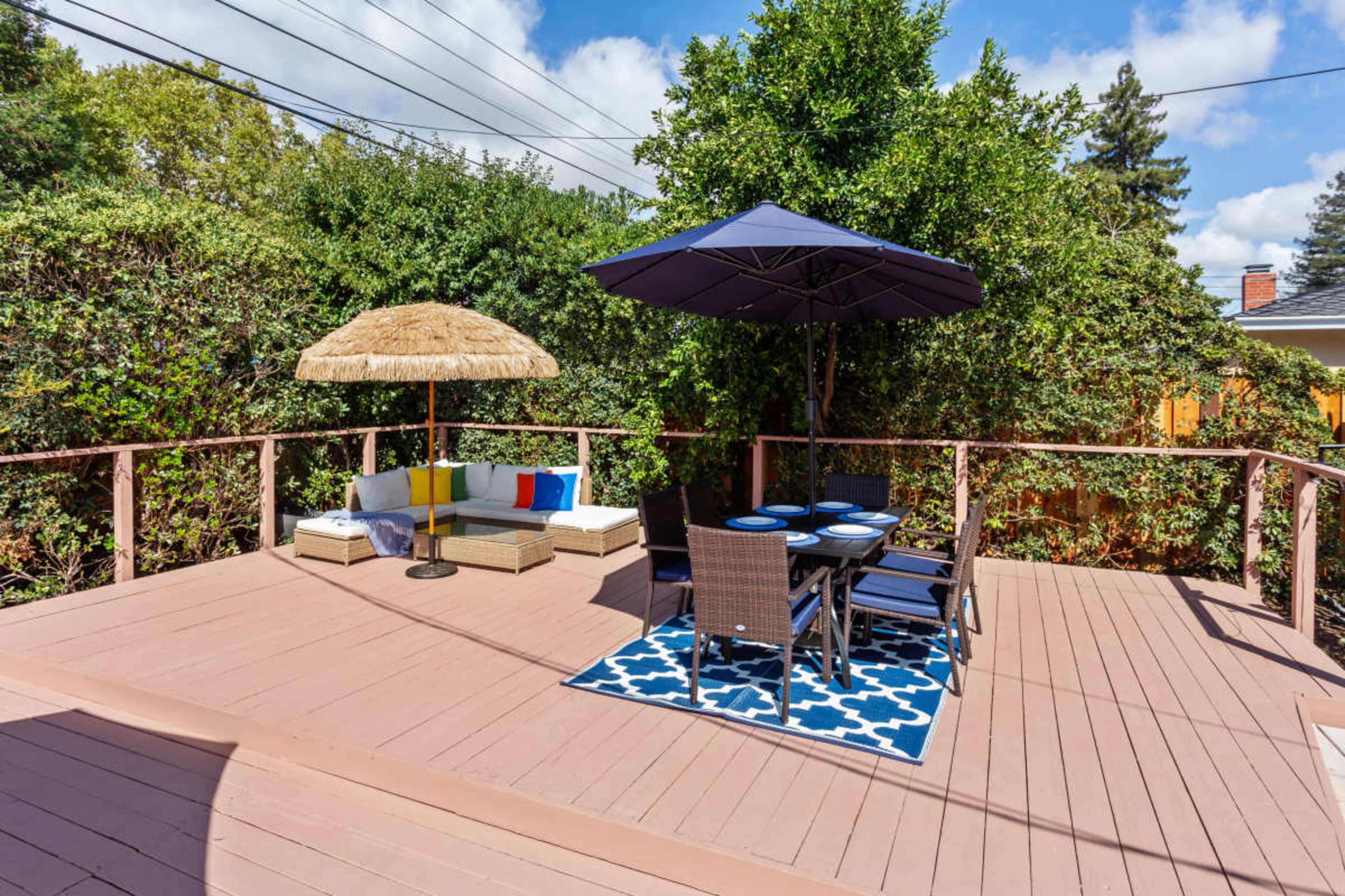 A wooden deck features a patio set with a table and chairs, a lounge area with cushions, and an umbrella beside a thatched sunshade.