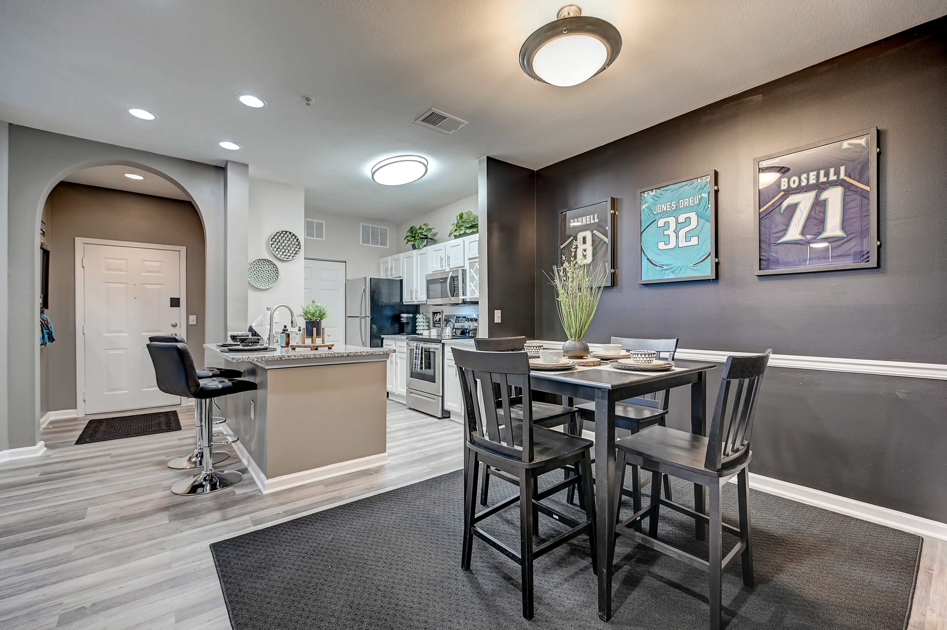 The image shows a modern kitchen and dining area featuring a black dining set, sports jerseys framed on the wall, and a well-equipped kitchen with white cabinets.